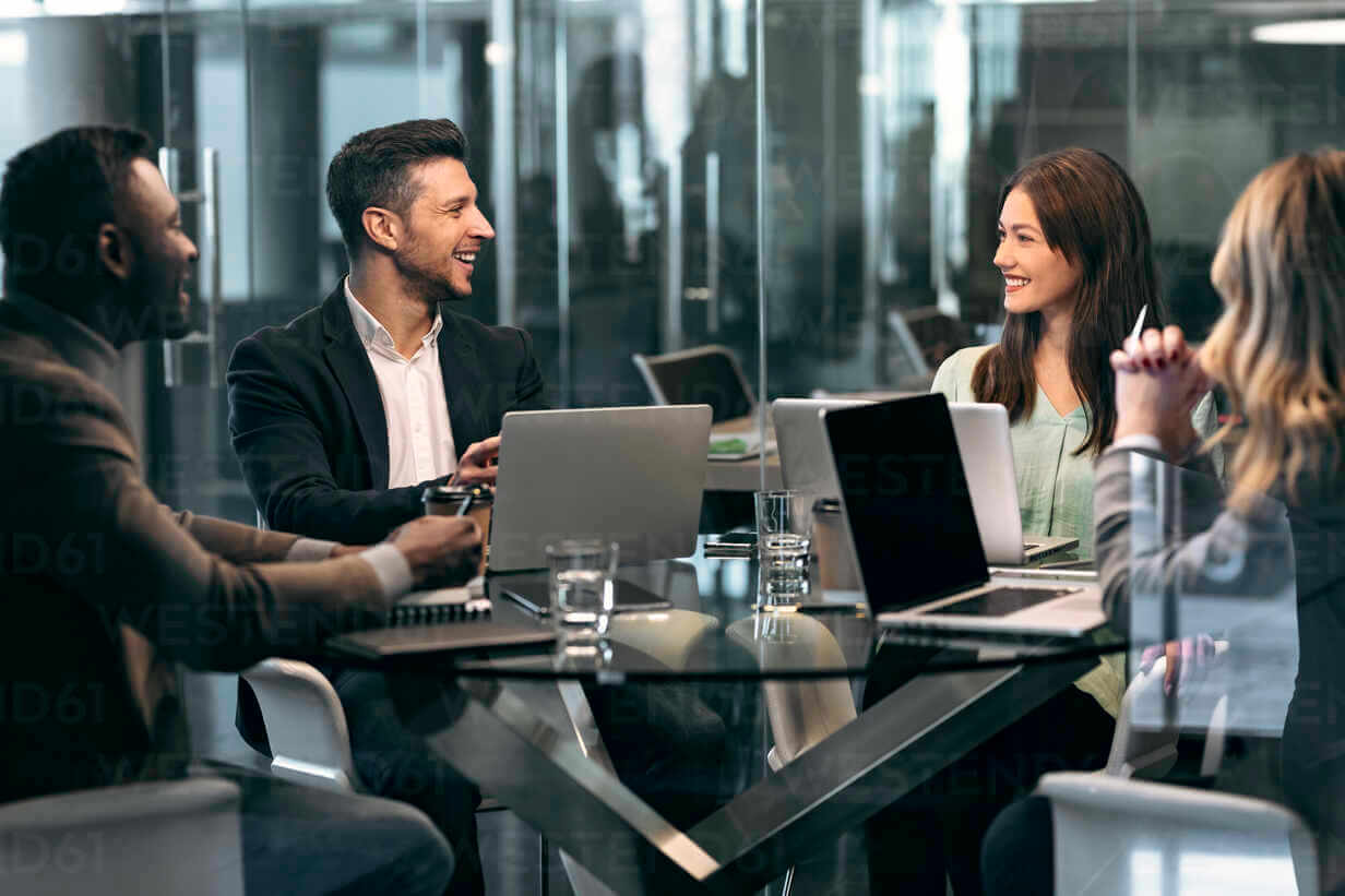 Four people sit around a glass table in a modern office, smiling and talking, with open laptops, notebooks, and glasses of water in front of them, suggesting a collaborative business meeting.