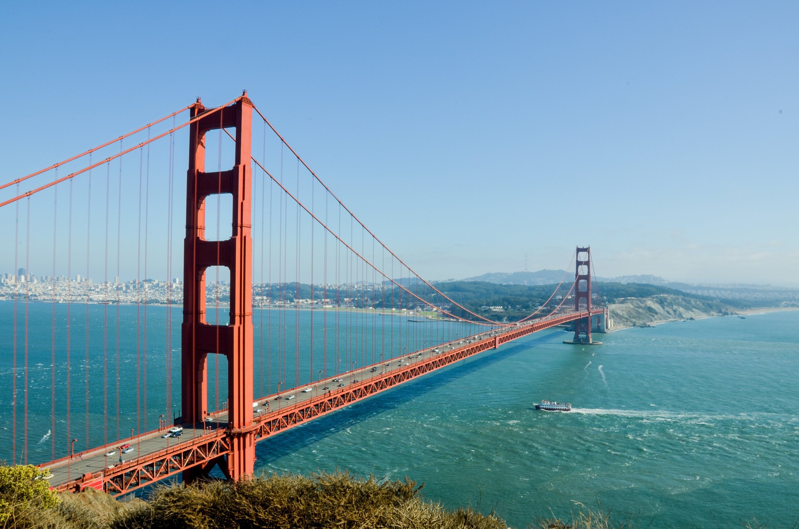 The Golden Gate Bridge spans across blue water under a clear sky, connecting San Francisco to Marin County. Cars cross the iconic red suspension bridge, and a boat sails below. Distant hills and cityscape are visible in the background.