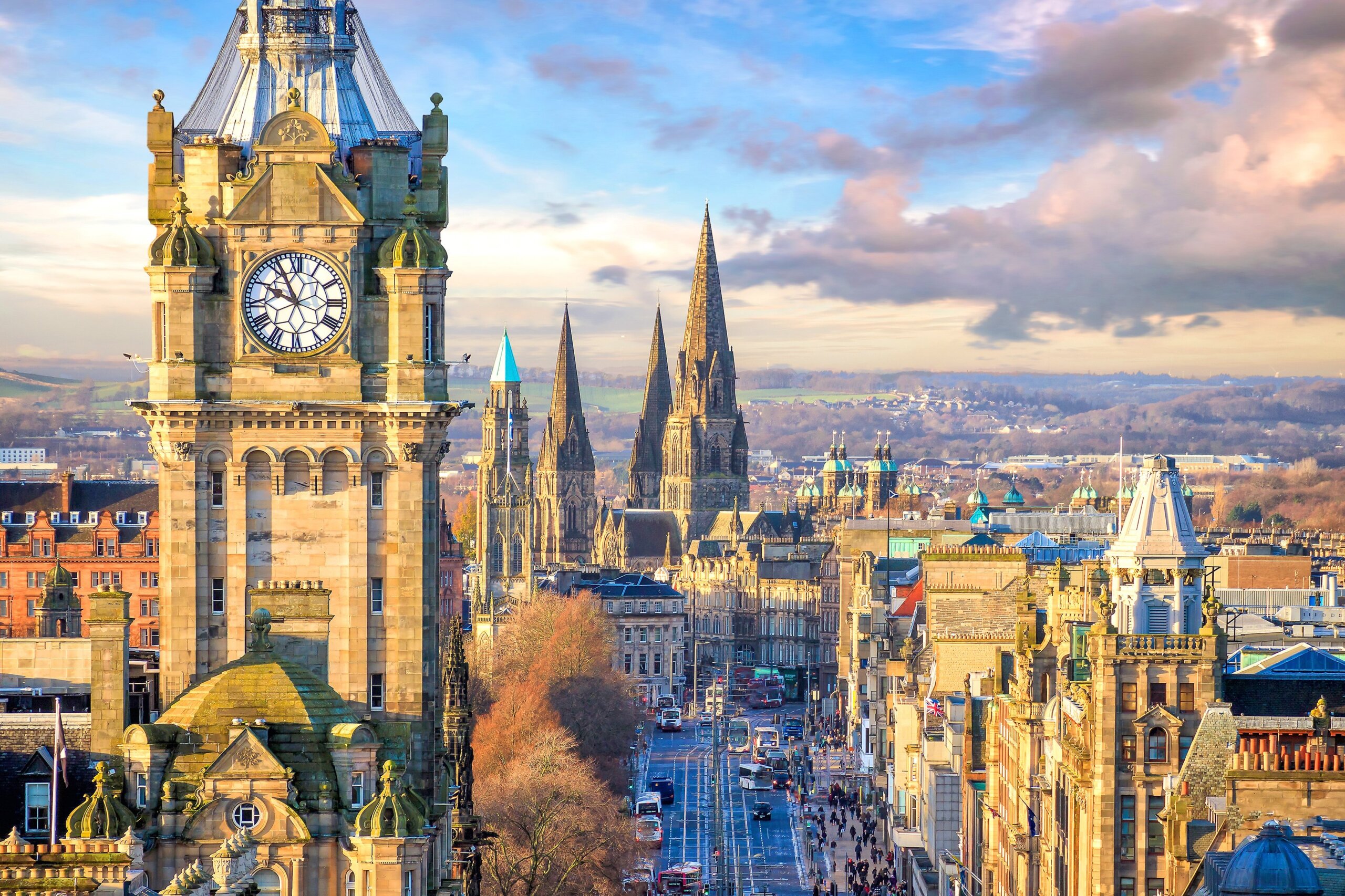 A scenic cityscape of Edinburgh, Scotland, featuring the Balmoral Hotel clock tower in the foreground, historic buildings, and church spires under a partly cloudy sky.