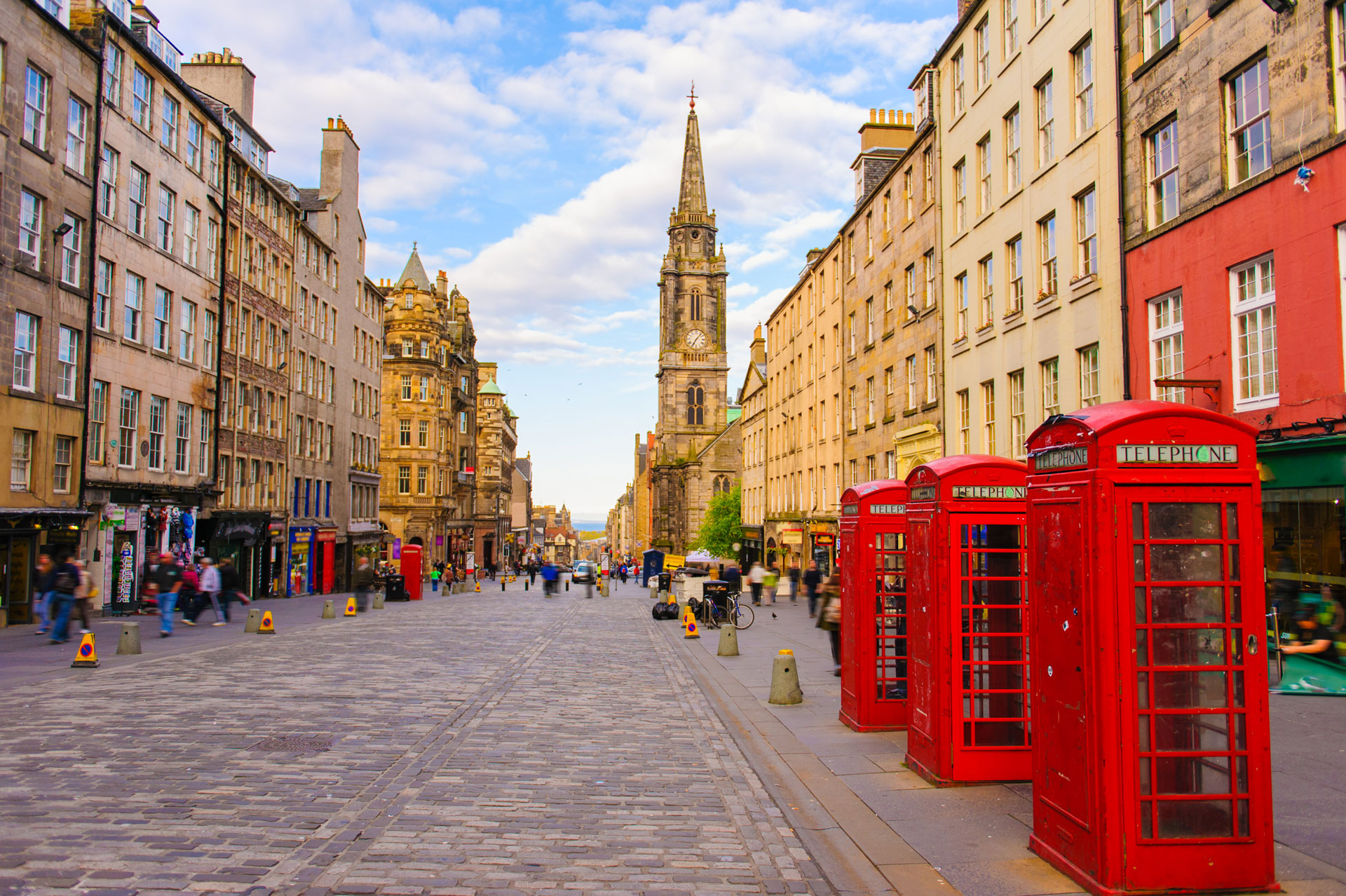 A cobblestone street lined with historic buildings and four red British phone booths, leading to a tall church spire under a partly cloudy sky, with people walking and shopping.