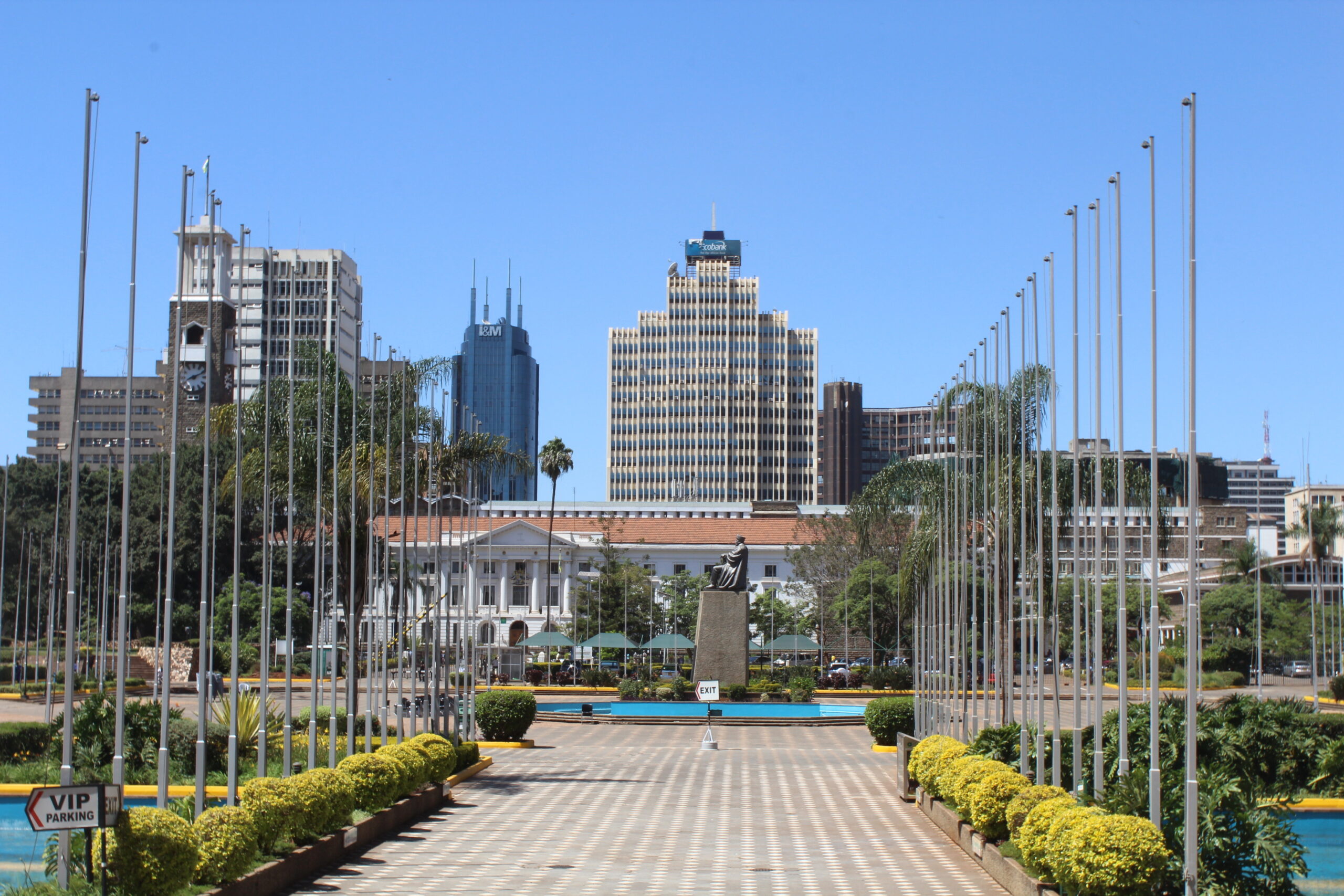 A pathway lined with flagpoles leads to a statue in front of a white building, with modern skyscrapers and clear blue sky in the background. Green shrubs border the walkway.