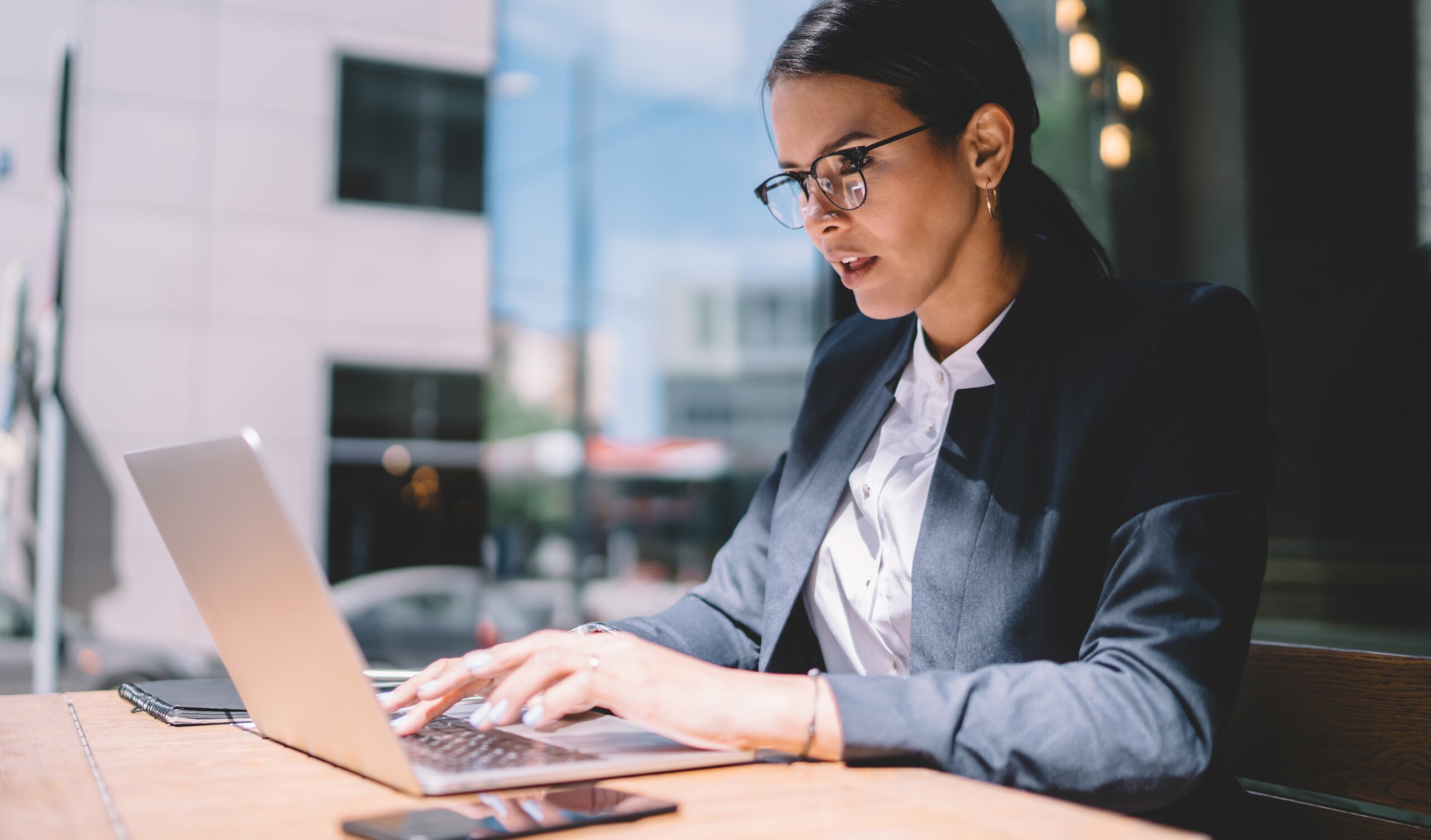 A woman wearing glasses and a business suit works on a laptop at an outdoor cafe table, with a smartphone and notebook beside her. City buildings and large windows are visible in the background.