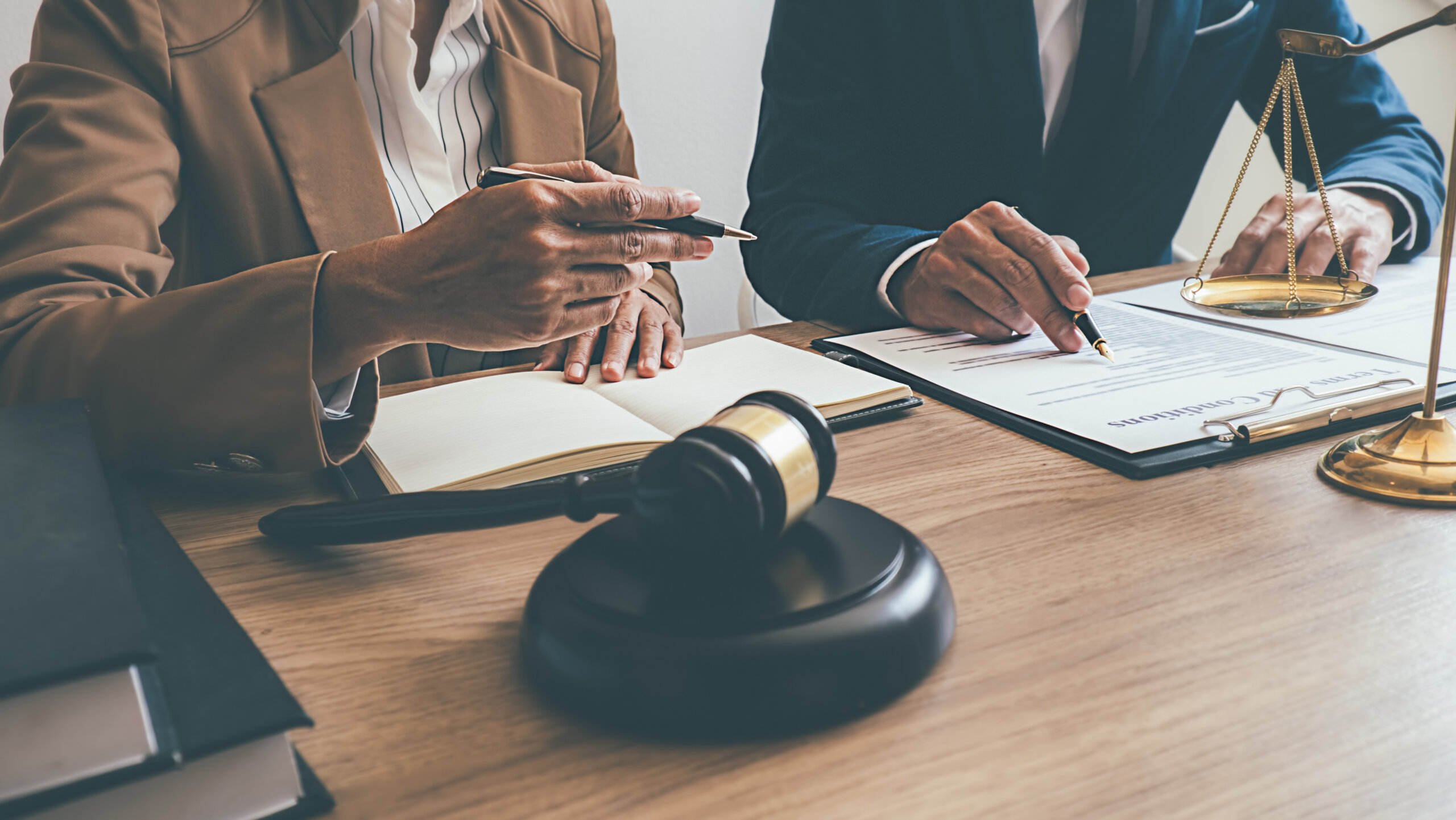 Two people in business attire sit at a desk with legal documents, a gavel, and scales of justice, discussing paperwork and making notes, suggesting a legal or professional consultation.