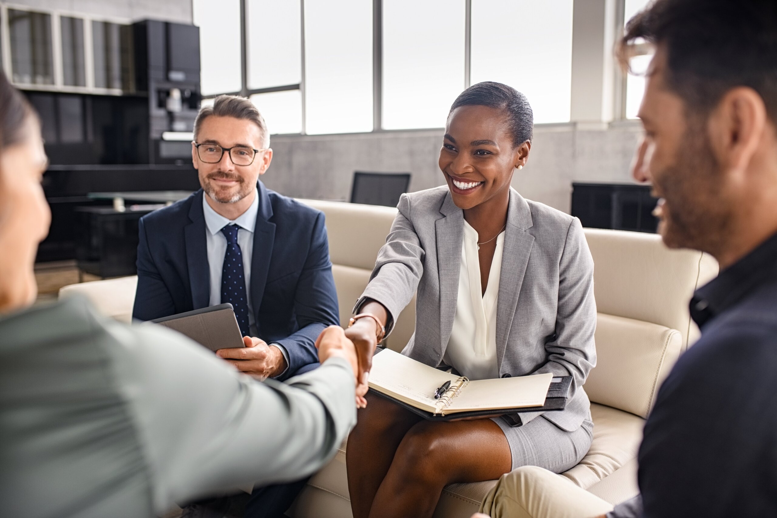 Four business professionals sit together in an office. A woman in a gray suit smiles while shaking hands with another person. Two men watch and smile. Papers and a tablet are visible, suggesting a meeting or interview.