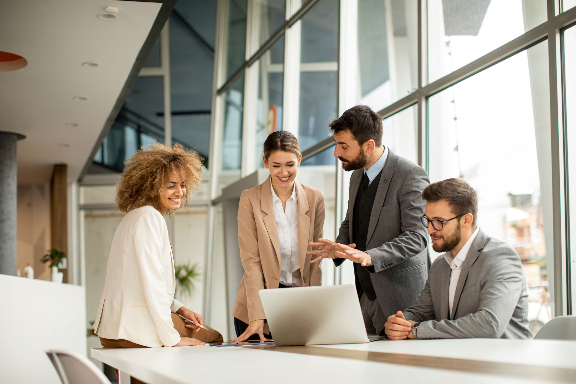 Four professionals in business attire stand and sit around a laptop in a modern office, discussing and smiling. Large glass windows let in natural light, creating a bright and collaborative atmosphere.