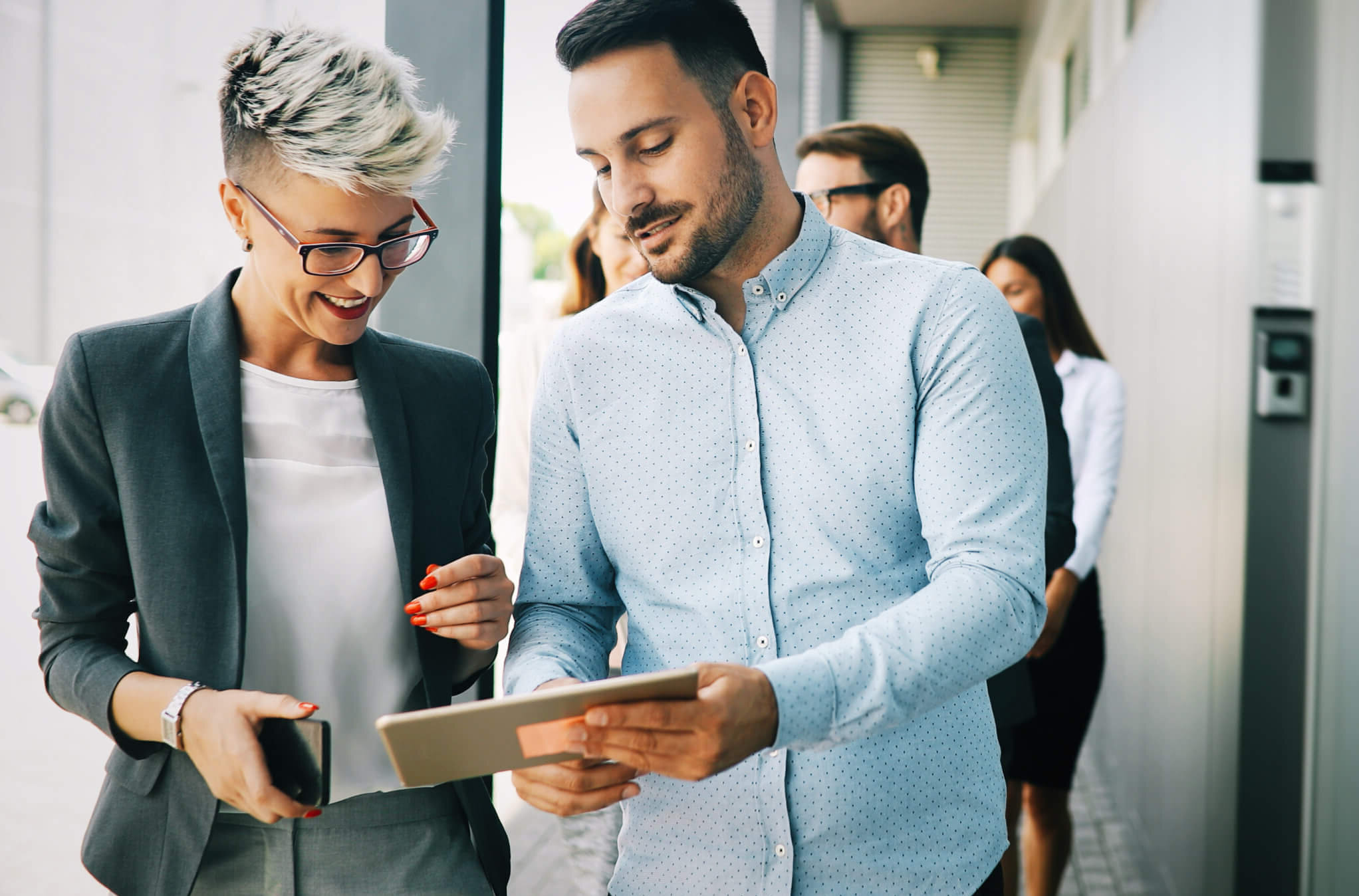 Two business professionals, a woman and a man, stand outside a modern building, smiling and discussing something on a digital tablet. Other people are visible in the background.