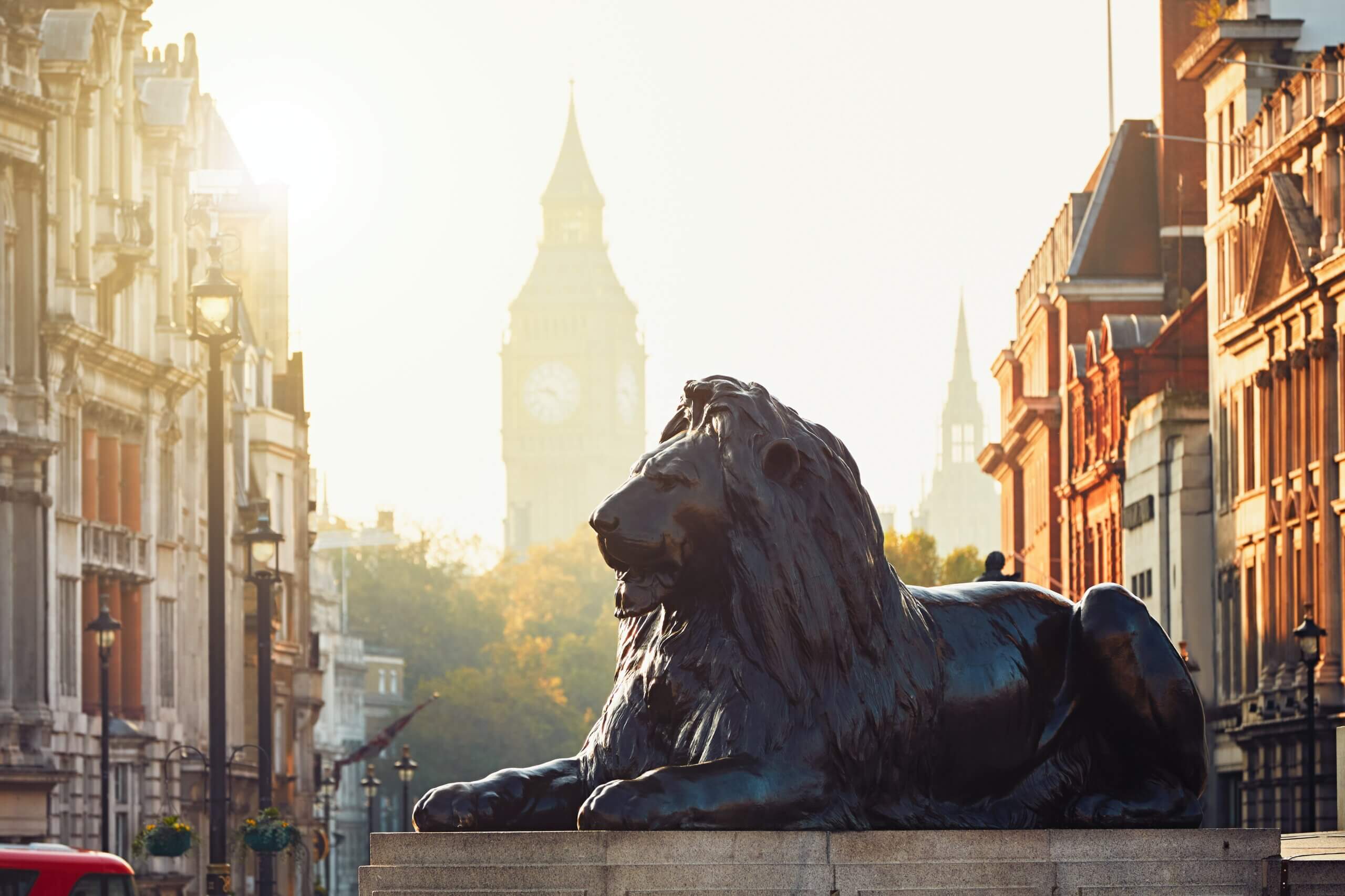 A bronze lion statue in Trafalgar Square, London, with historic buildings lining the street and the clock tower of Big Ben visible in the hazy background at sunrise.