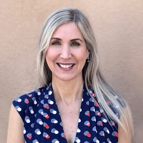 A woman with long, straight blonde hair is smiling at the camera. She is wearing a navy blue sleeveless top with a red and white polka dot pattern, standing in front of a plain beige background, perhaps preparing for an intellectual property law seminar.