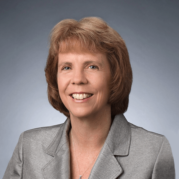 A woman with short, light brown hair smiles at the camera. She is wearing a light gray blazer over a light top and is posed against a plain, muted blue-gray background.