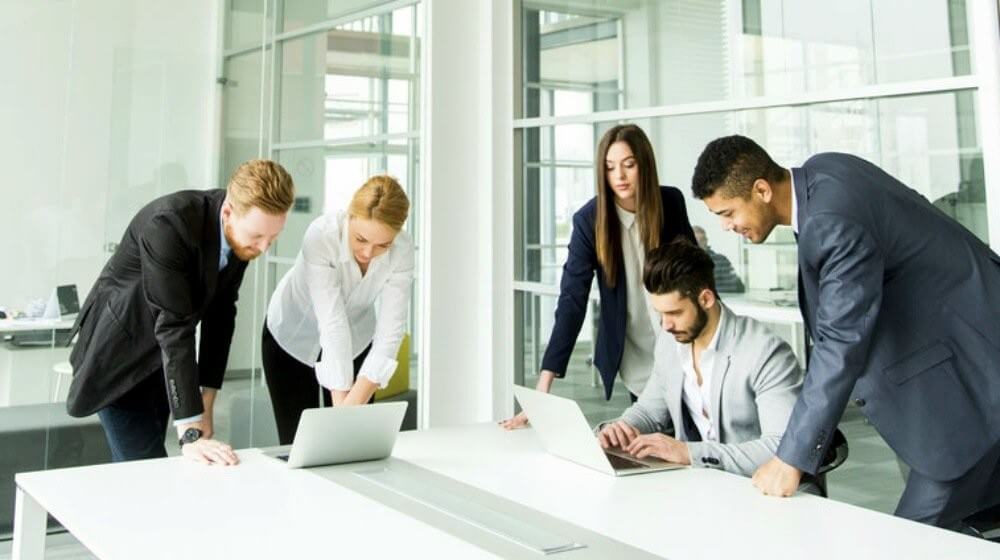 Five business professionals, three standing and two sitting, collaborate in a bright, modern office. They are focused on laptops and documents on a white table, engaged in discussion. Large windows let in natural light.