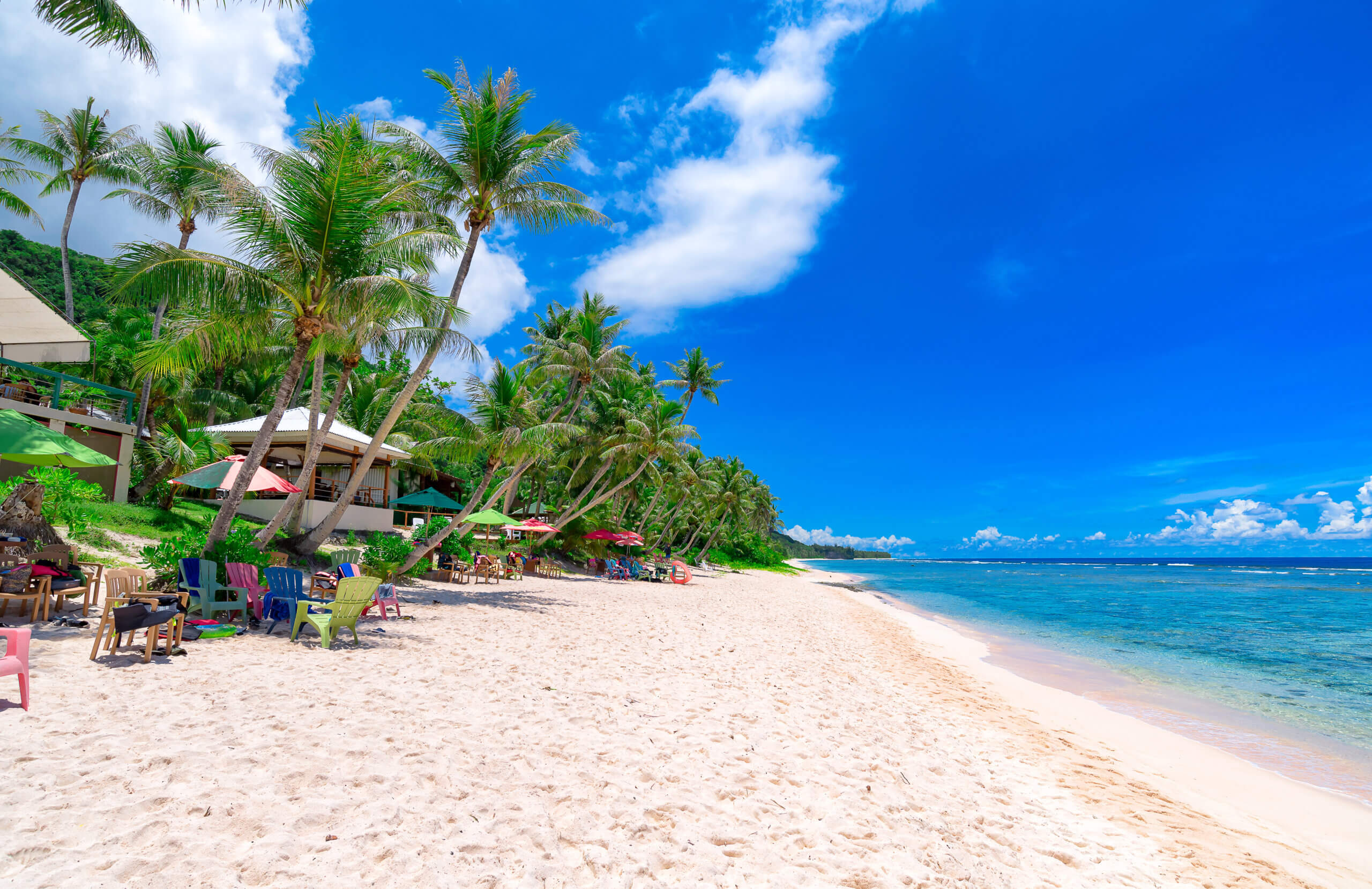 A tropical beach with white sand, clear blue water, palm trees, colorful chairs, and sun umbrellas under a bright, sunny sky. Lounge areas and greenery are visible along the left side.