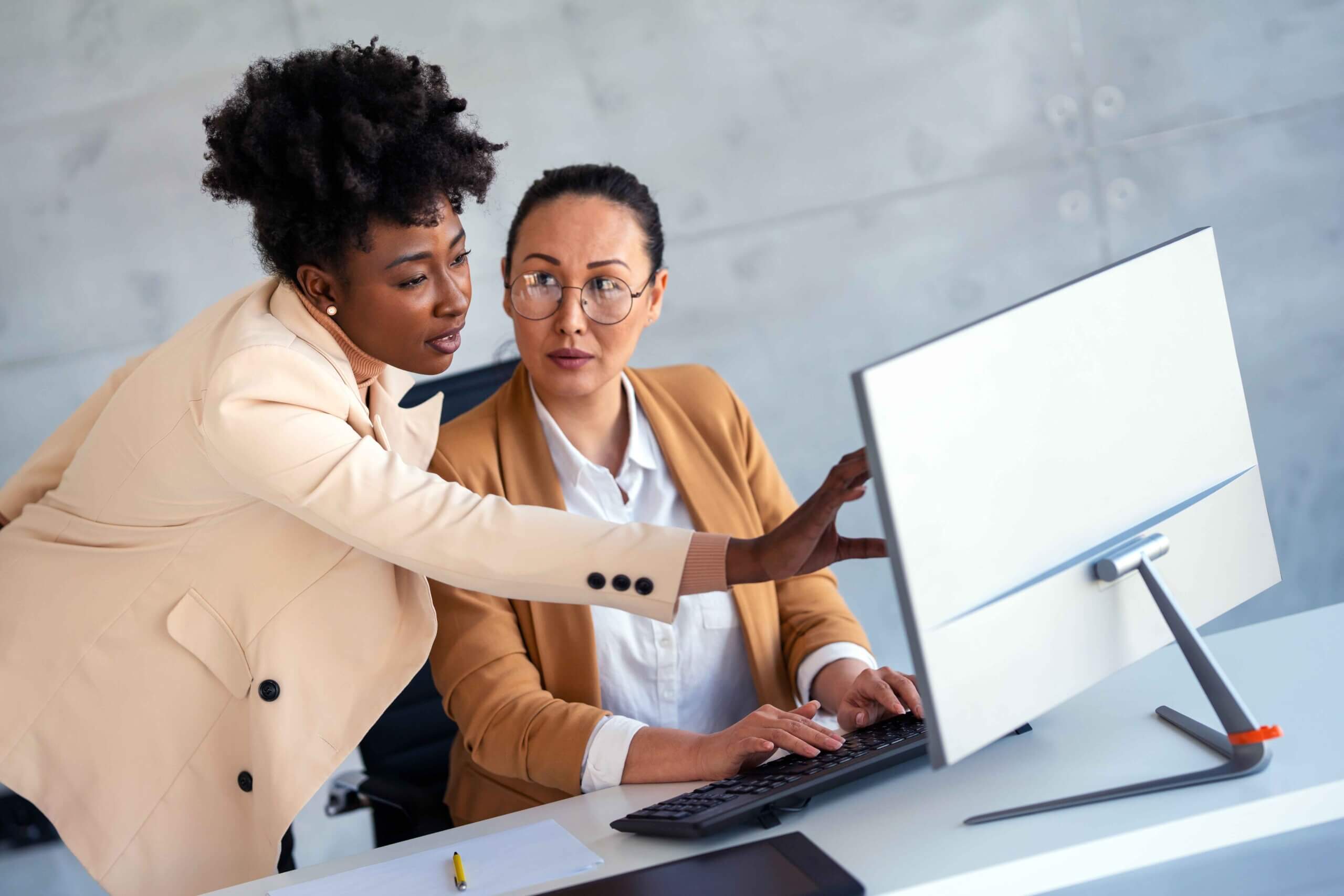 Two women in business attire work together at a desk. One woman is seated using a computer, while the other stands beside her, pointing at the monitor, both focused on the screen in an office setting.