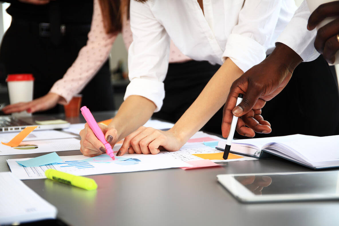 Several people collaborate at a desk, pointing at documents and charts with pens and highlighters. Their focus is on paperwork spread across the table, suggesting a teamwork or business planning session.