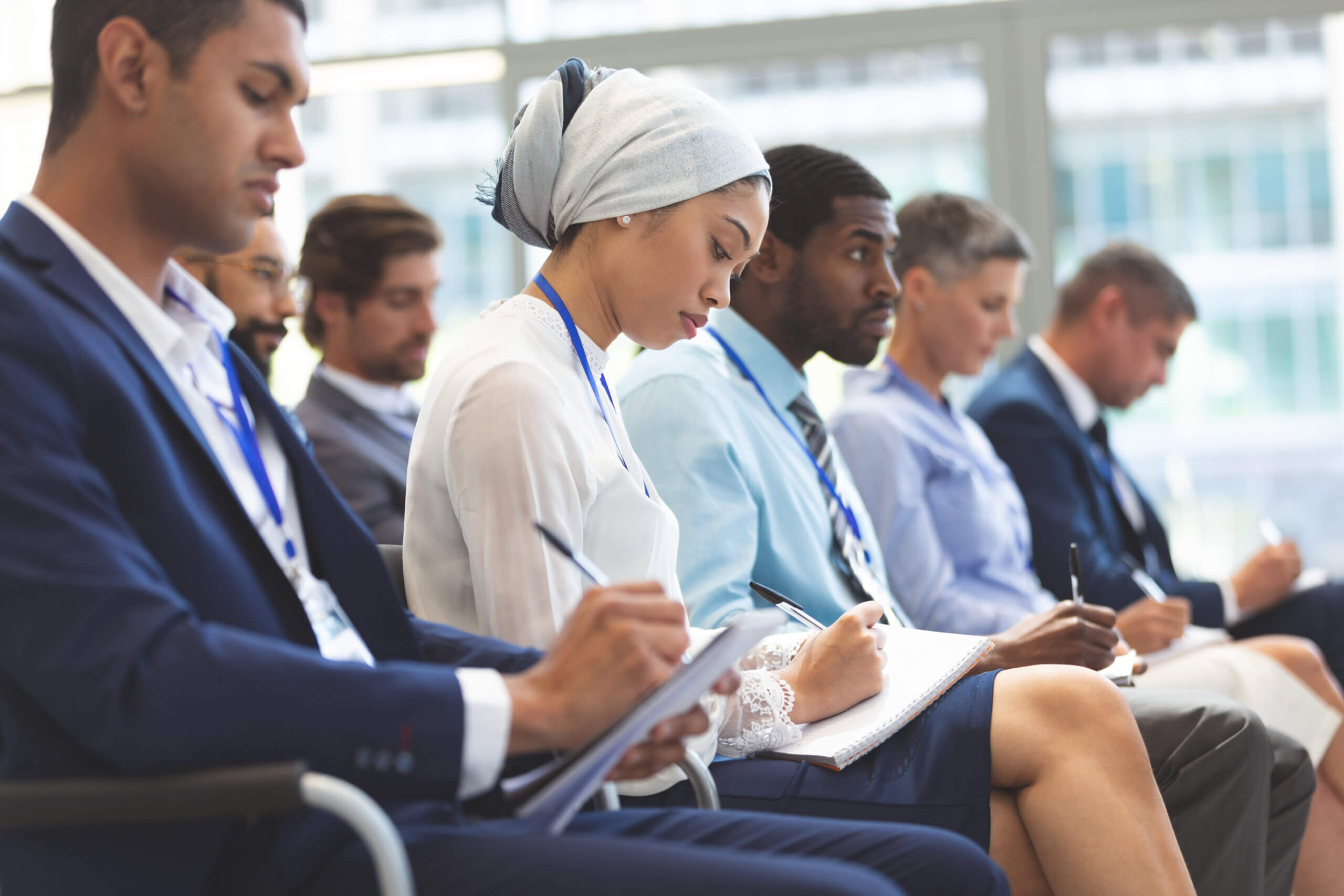 A diverse group of professionals sits in a row at a conference or seminar, attentively taking notes on notepads. They are wearing formal attire and conference lanyards, with large windows in the background.