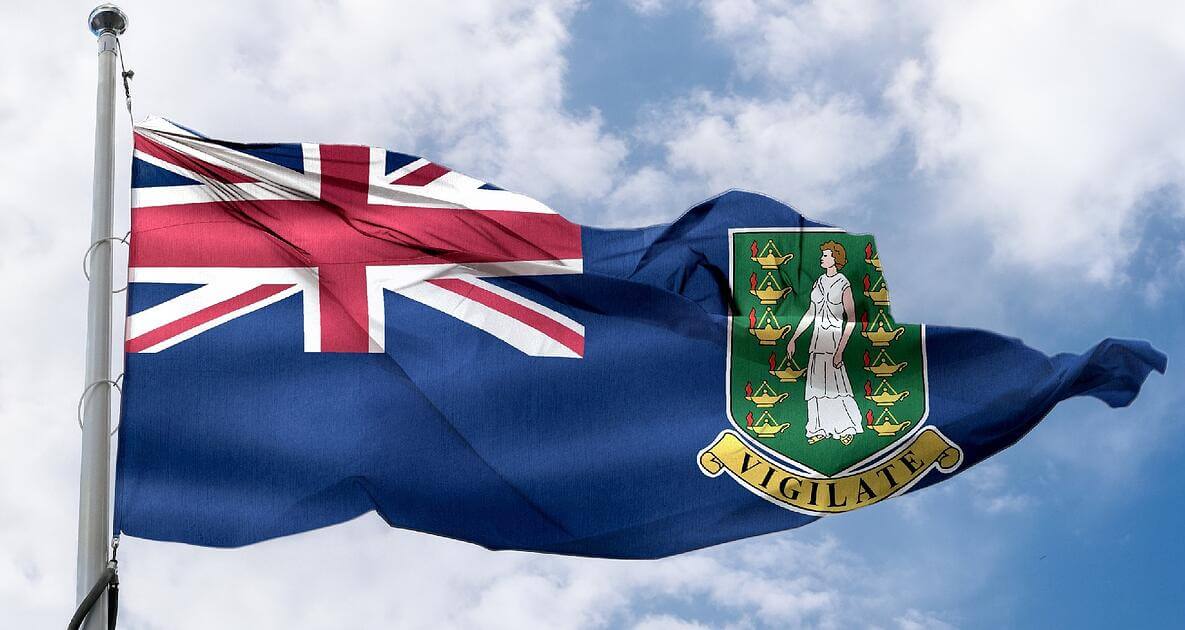 The flag of the British Virgin Islands waving against a blue sky, featuring a Union Jack in the top left corner and the territory’s coat of arms with a woman and several lamps on the right side.