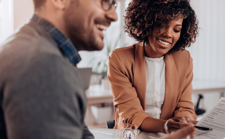 Two people sit together at a table, smiling and looking at documents. The woman holds a pen and wears a tan blazer, while the man wears glasses and a grey sweater. They appear to be having a pleasant conversation.