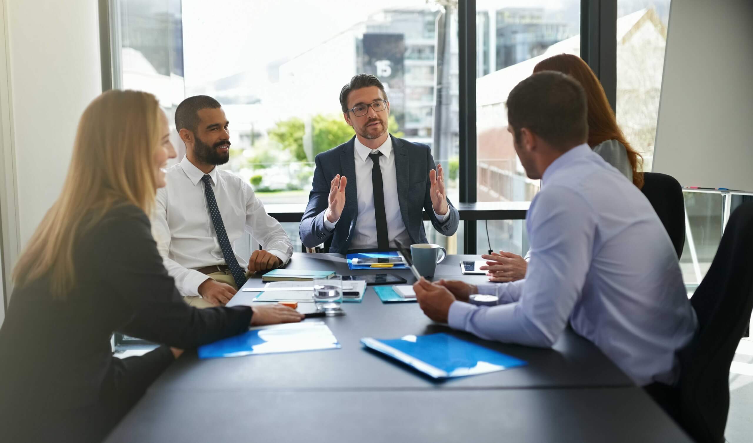 A group of six professionally dressed people sit around a conference table in a modern office, engaged in a discussion. Papers, folders, and coffee cups are on the table, with city buildings visible through large windows.