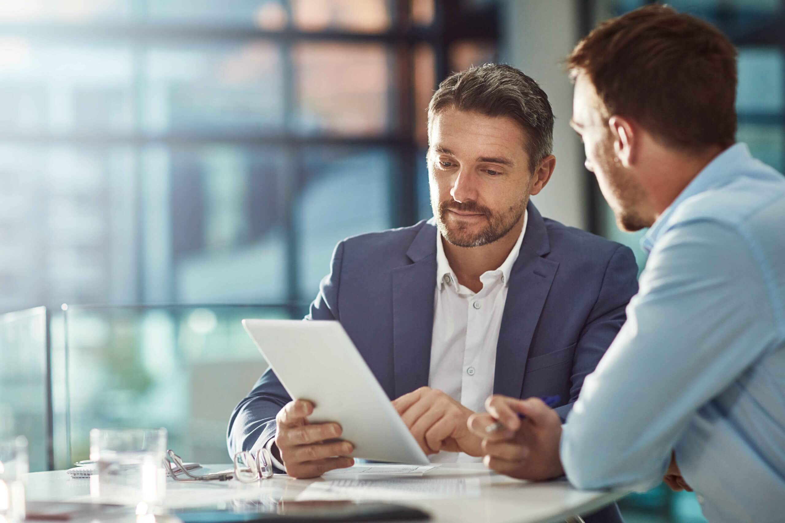 Two men in business attire sit at a table in a modern office, one holding a digital tablet and showing it to the other as they have a discussion. Sunlight streams through large windows in the background.