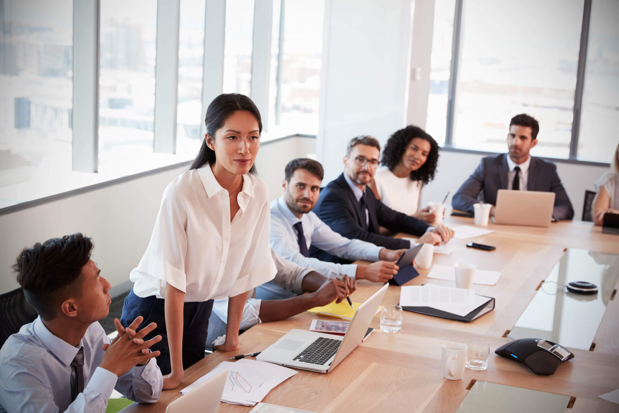 A woman stands at a conference table, presenting to a group of colleagues who are seated and listening, with laptops, papers, and coffee cups on the table in a modern office setting.
