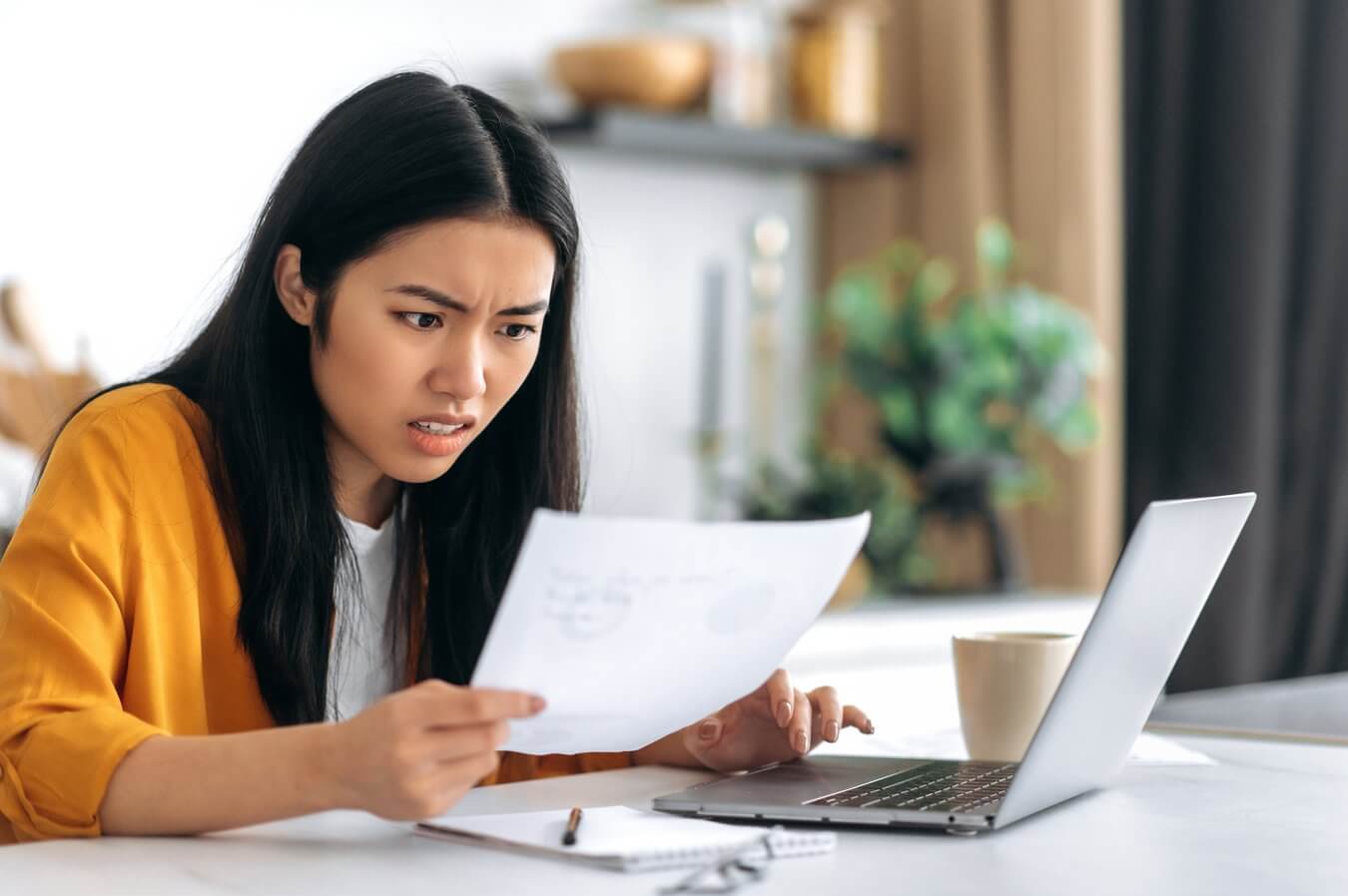 A woman sitting at a desk looks concerned while reading a piece of paper. She has a laptop, notebook, pen, and coffee cup in front of her, and appears focused on the document.