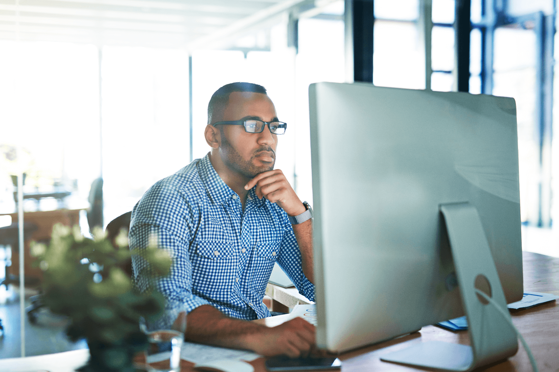 A man wearing glasses and a checkered shirt sits at a desk, looking thoughtfully at a large computer monitor in a bright modern office.