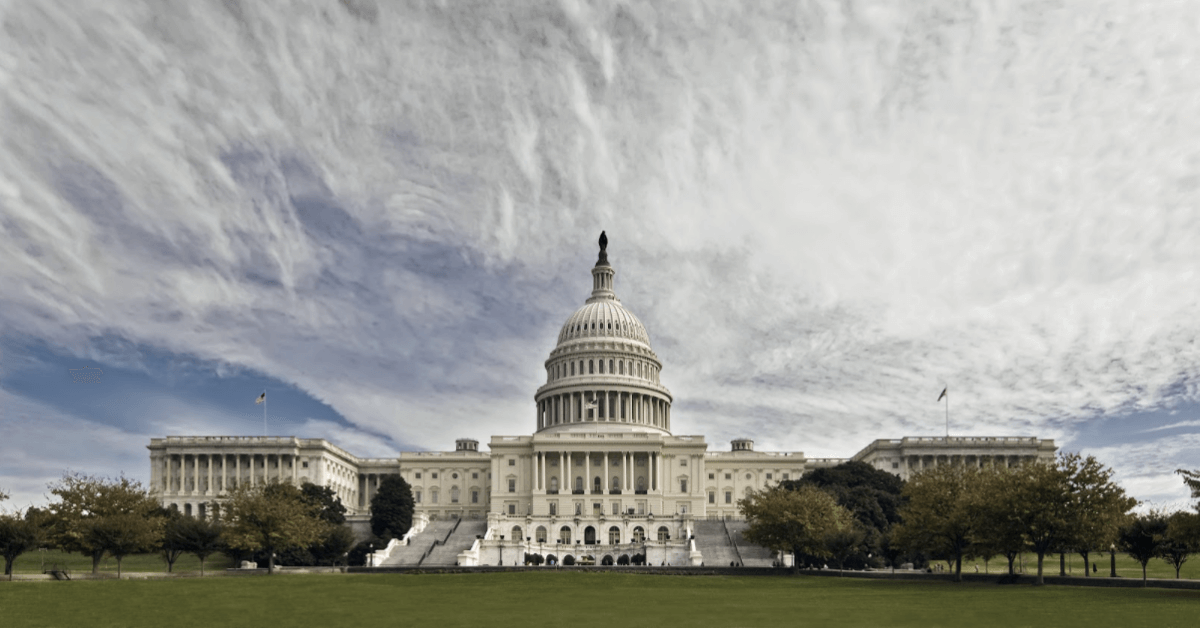 The United States Capitol building with its white dome, surrounded by trees and a green lawn under a dramatic, cloudy sky.