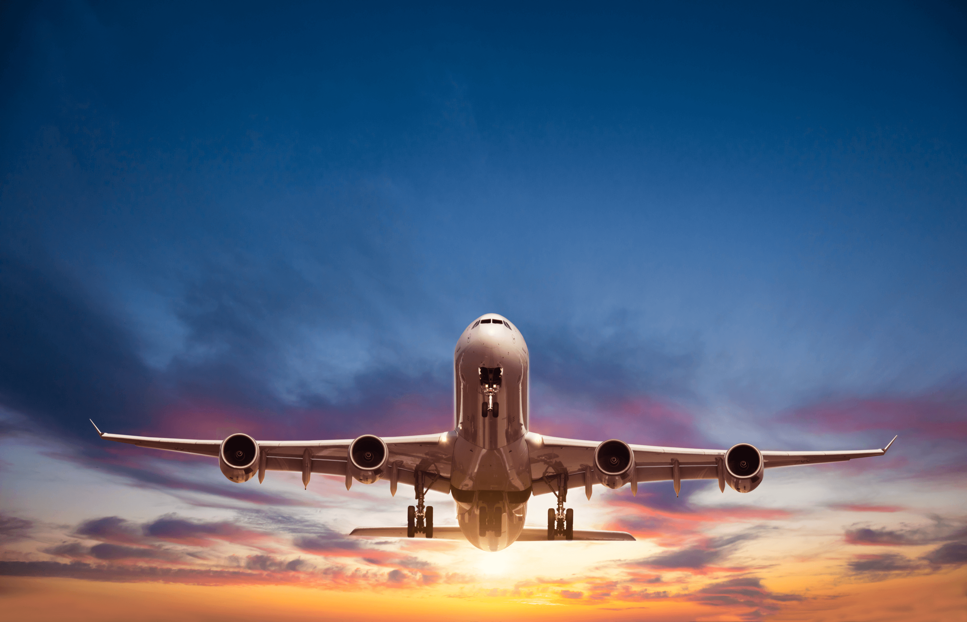 A large airplane flying overhead at sunset, with its landing gear extended and a colorful sky of blue, pink, and orange hues in the background.