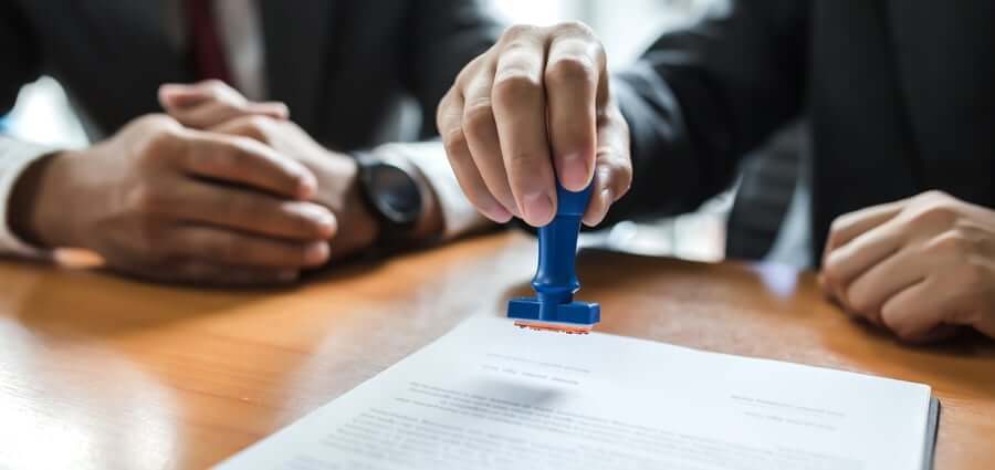Two people in business attire sit at a desk; one person uses a blue stamp to approve or authorize a document on the table.