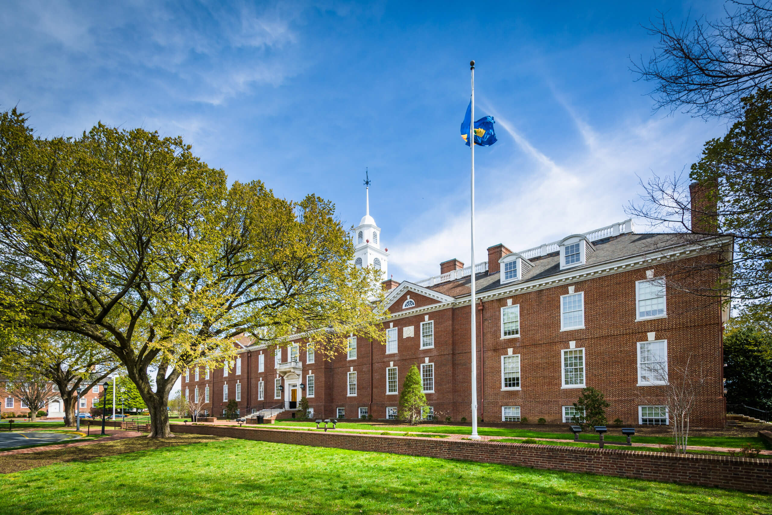 A red-brick government building with white trim and a central cupola, set behind a green lawn and large trees, with a blue state flag flying on a tall flagpole under a bright, partly cloudy sky.