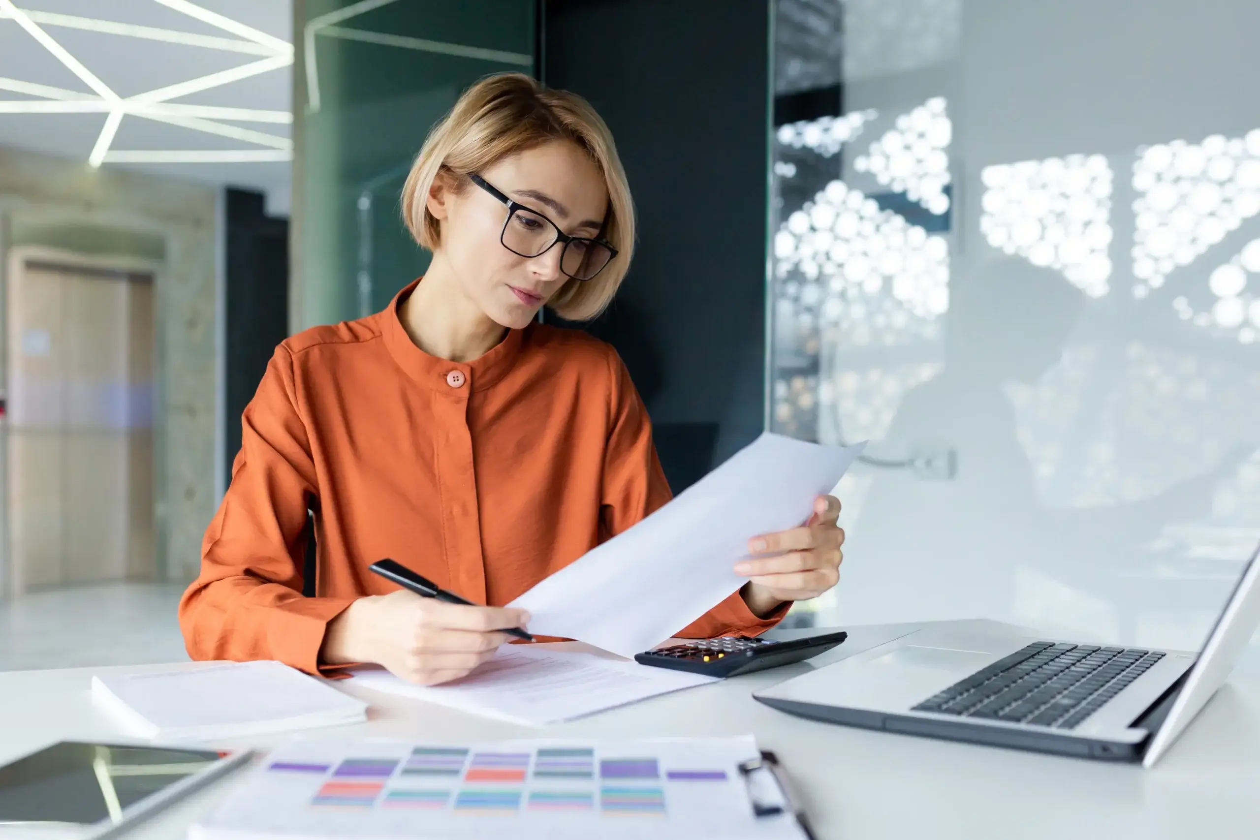 A woman wearing glasses and an orange blouse sits at a desk, reviewing documents and writing notes. A laptop, calculator, and colorful chart are on the table in front of her.