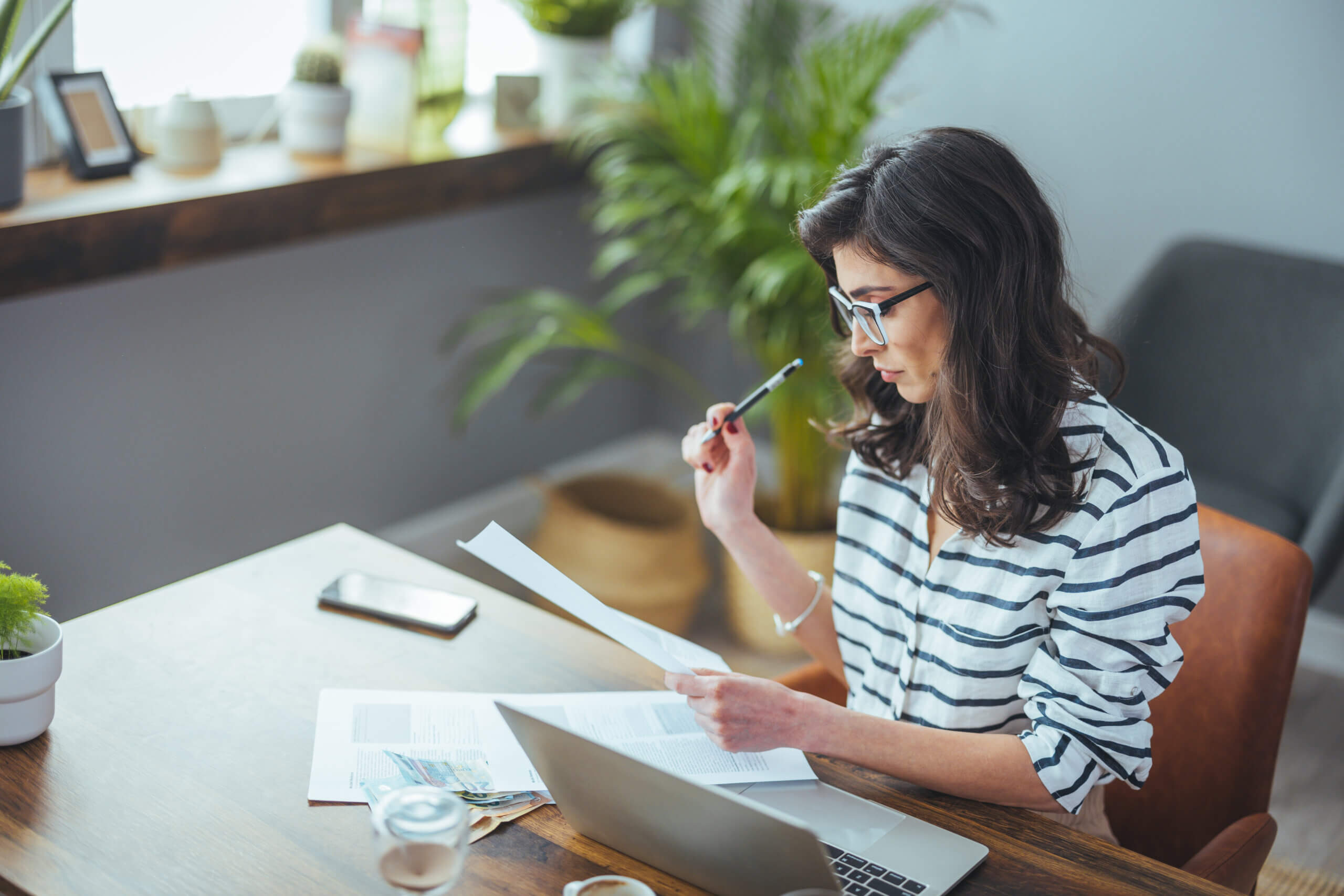 A woman wearing glasses sits at a desk with a laptop, papers, and a cup of coffee. She holds a pen to her chin and reads a document, surrounded by plants and natural light.