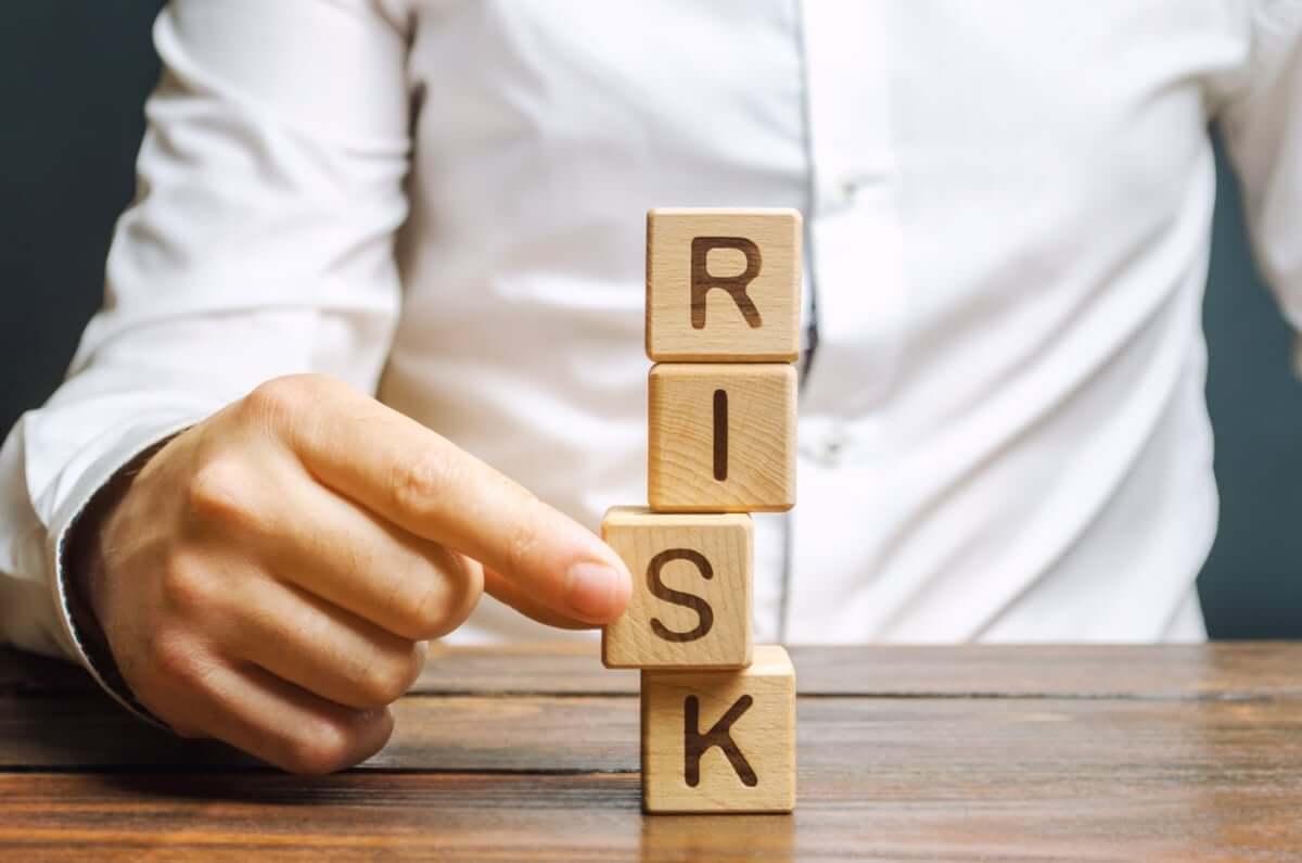 A person in a white shirt stacks wooden blocks spelling "RISK" on a wooden table, with one hand about to remove the "S" block.