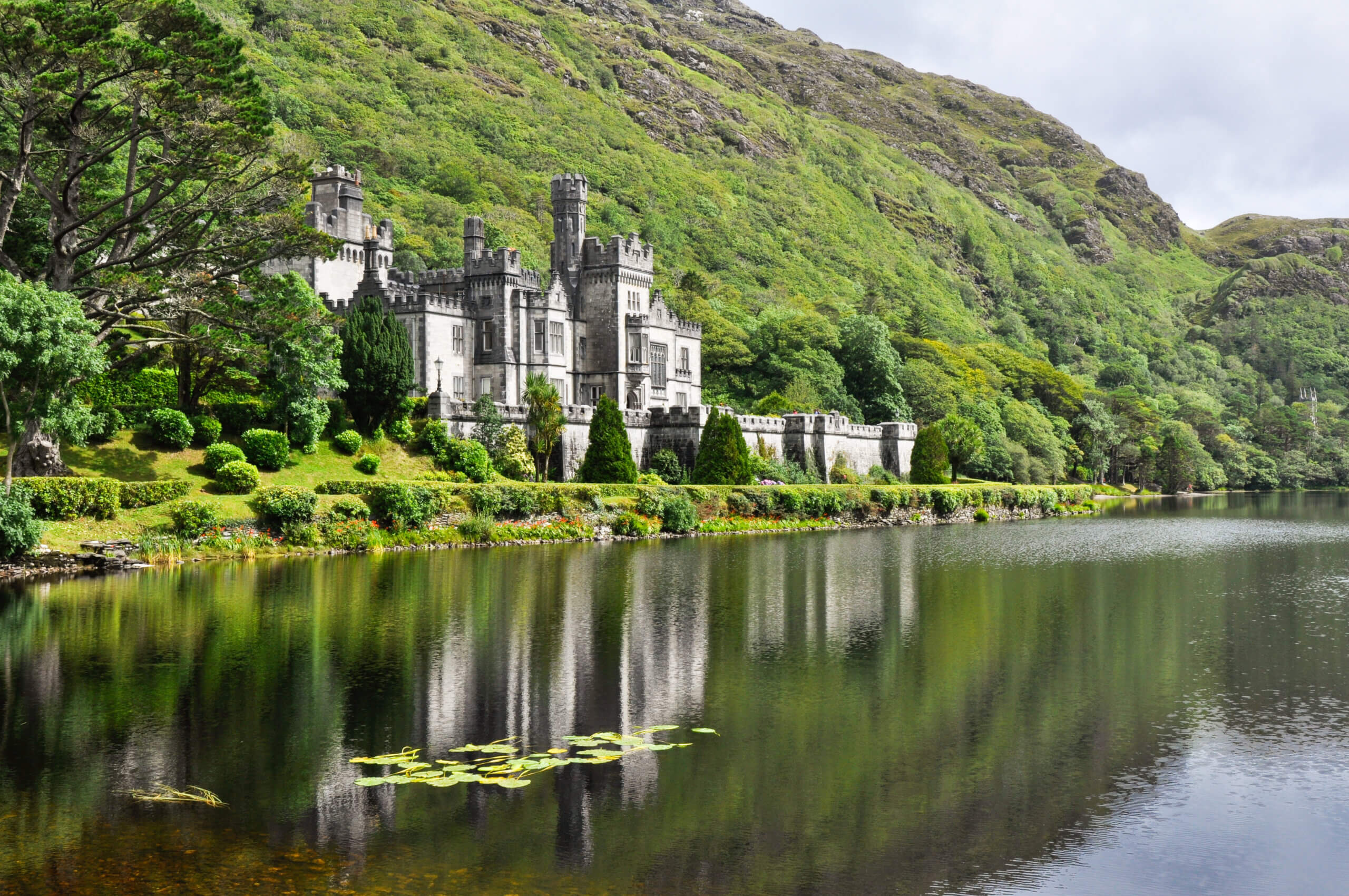 A large stone castle with turrets sits beside a calm lake, surrounded by lush green trees and hills, with the castle and landscape reflected in the water.