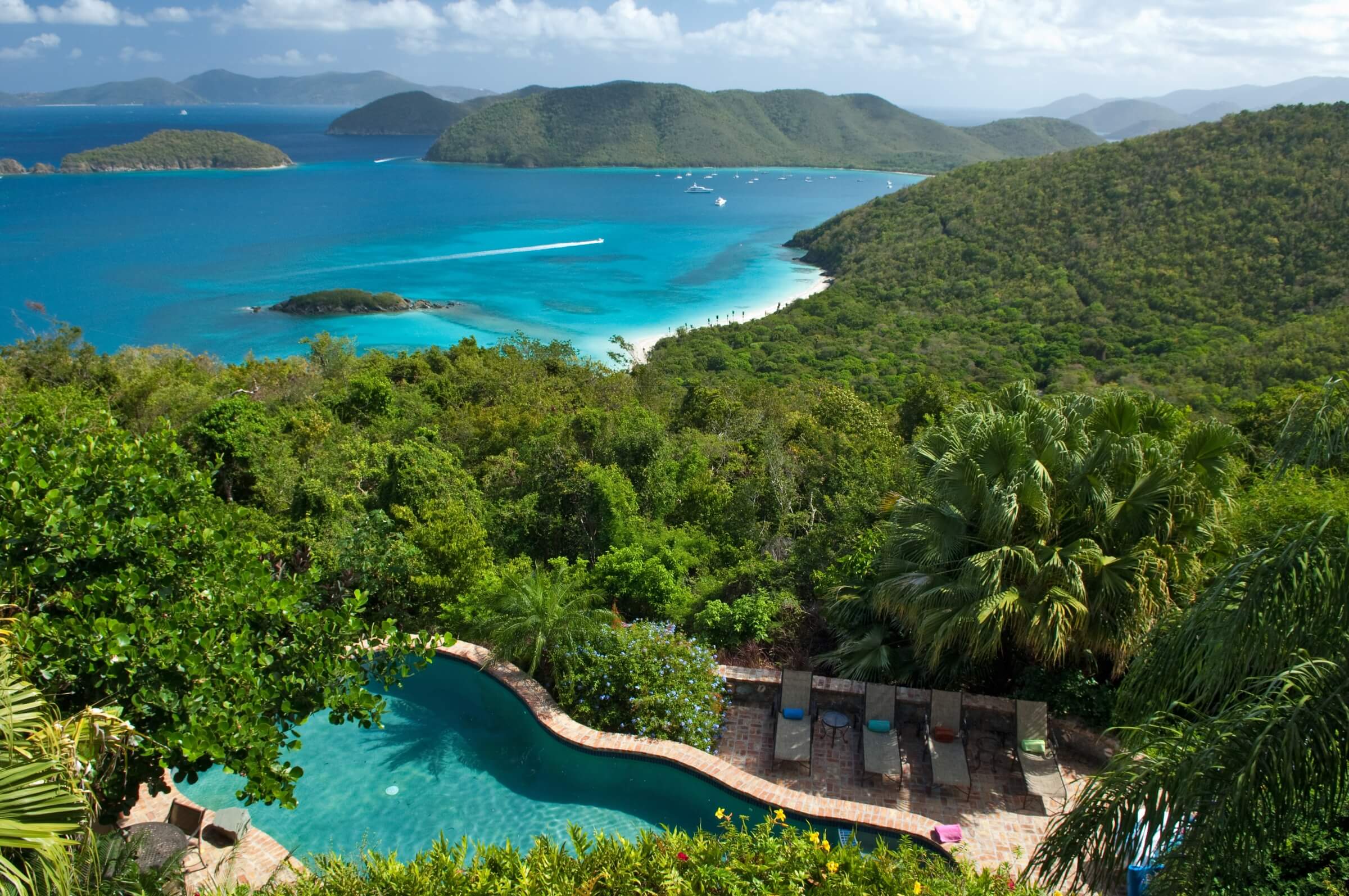 A tropical hillside overlooks a curved pool surrounded by palm trees, with turquoise ocean water, small islands, sandy beaches, and green hills in the background under a partly cloudy sky.