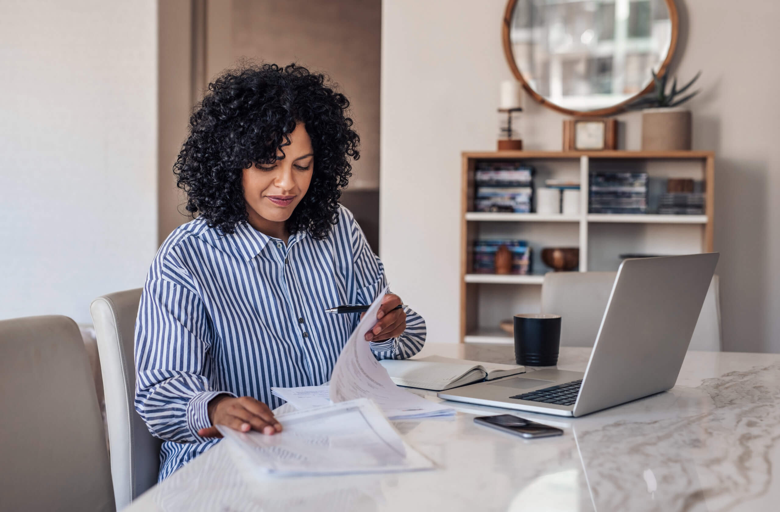 A woman with curly hair sits at a table with a laptop, pen, papers, and a mug, appearing focused as she reviews documents in a well-lit, modern home office.