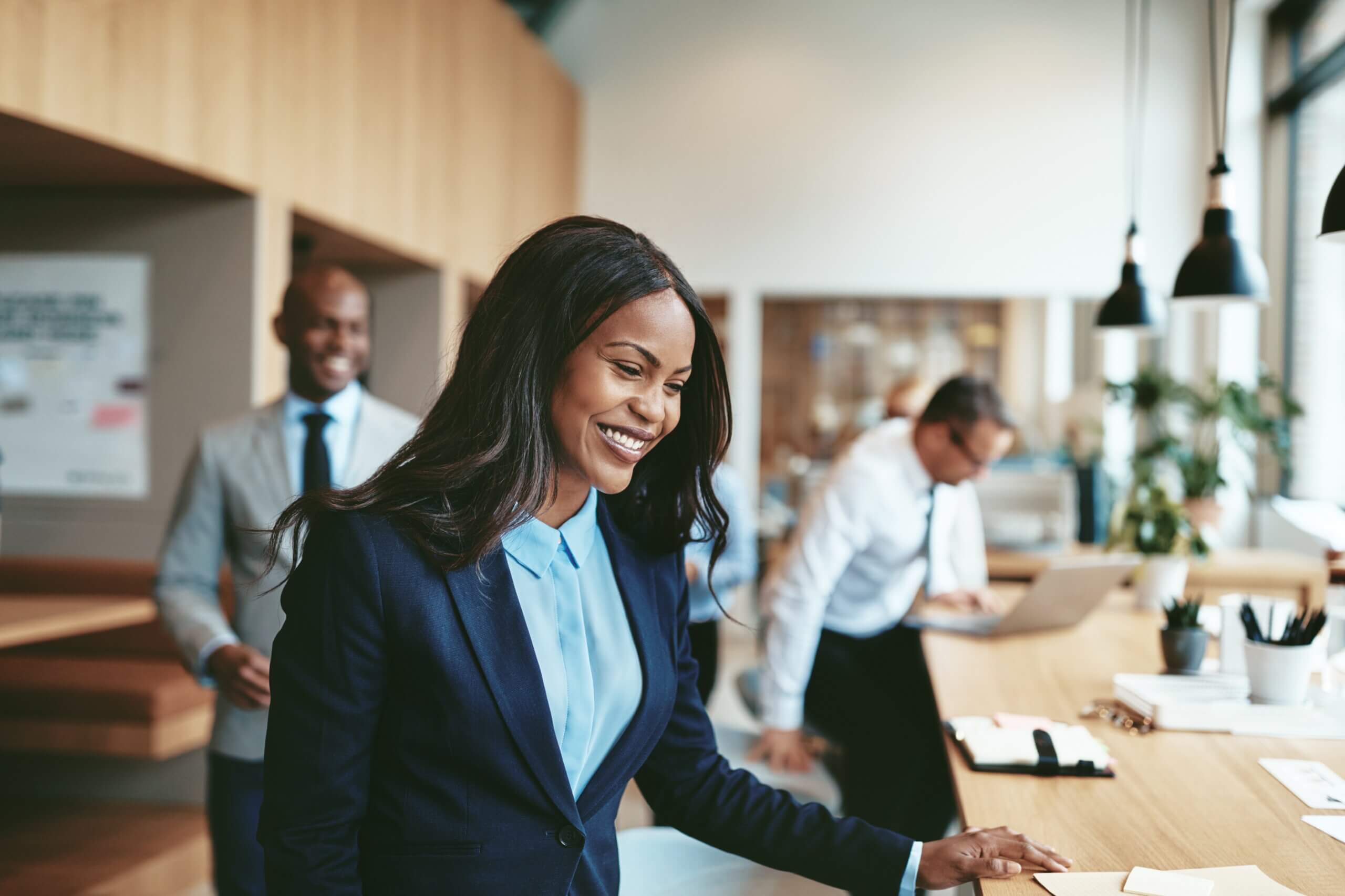 A smiling woman in a blue suit stands at a desk in a modern office, with two colleagues working in the background. Large windows let in natural light, creating a bright, professional atmosphere.