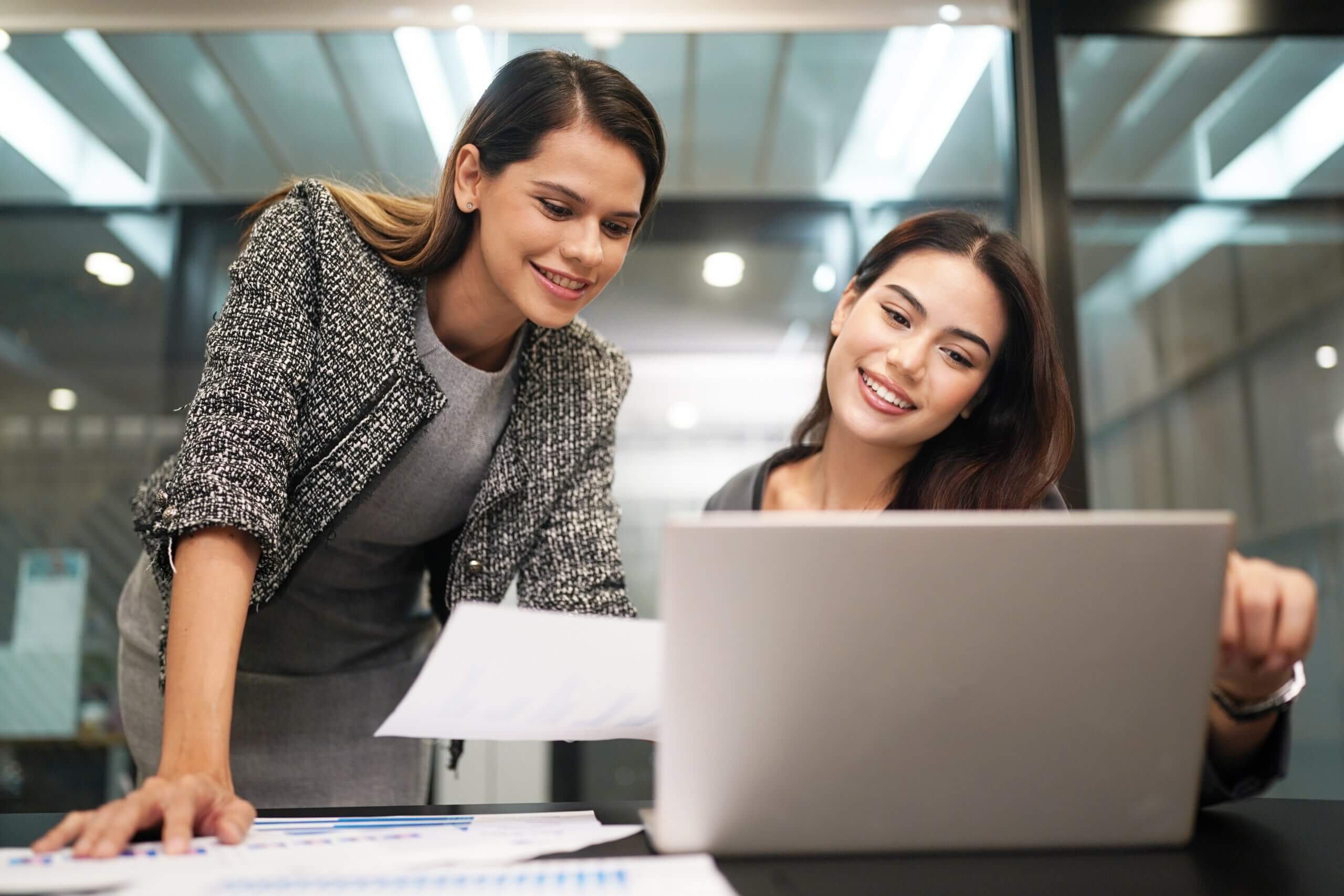 Two women in business attire smiling while looking at a laptop screen in an office. One is holding papers, and the desk has documents and charts spread out. They appear to be collaborating on a project.