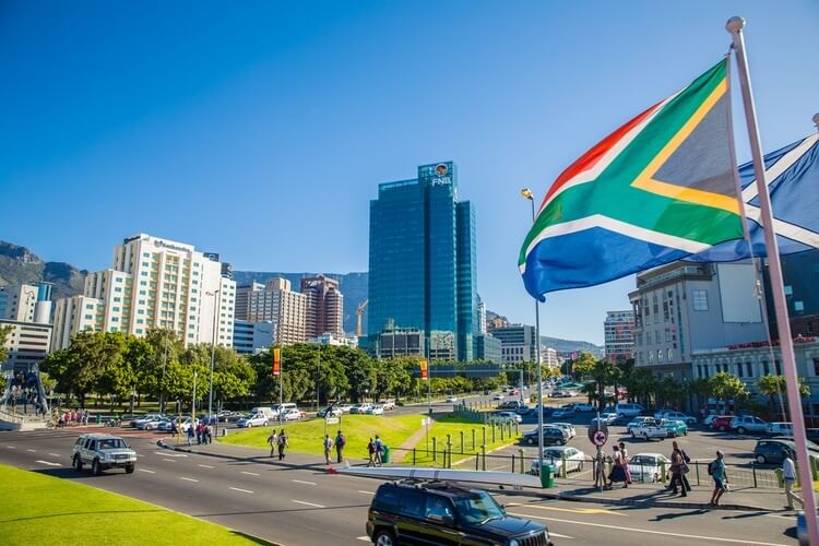 A busy city scene in Cape Town, South Africa, with cars on the road, people walking, high-rise buildings, and a large South African flag waving in the foreground on a sunny day.