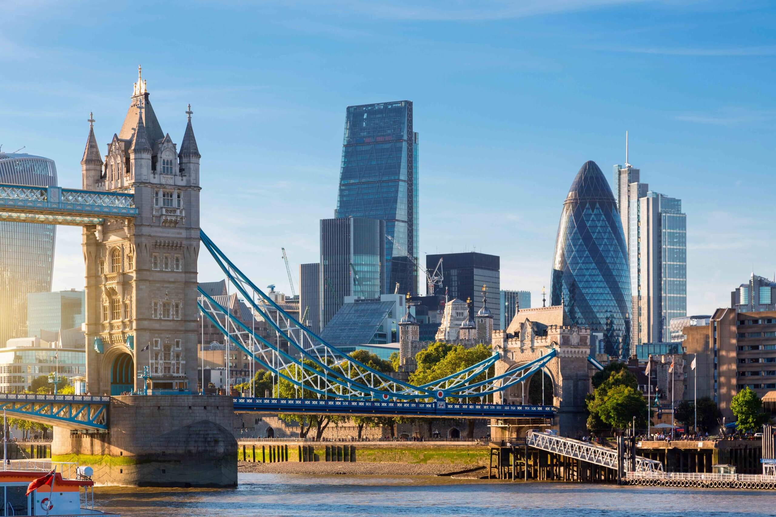 The image shows London’s skyline with Tower Bridge in the foreground and modern skyscrapers, including The Gherkin and The Leadenhall Building, in the background under a blue sky.