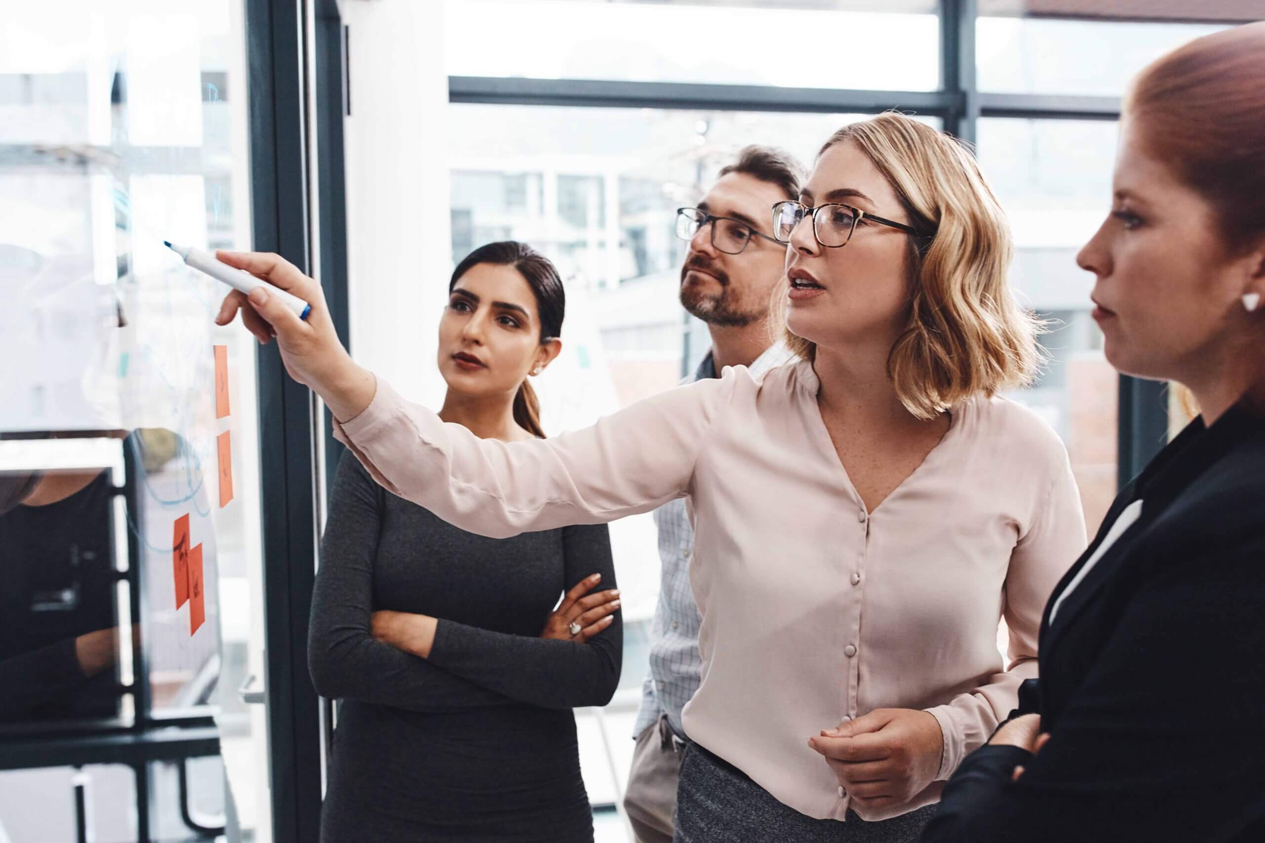 A woman in glasses is writing on a glass board with sticky notes while three colleagues attentively watch and listen during a meeting in a modern office.