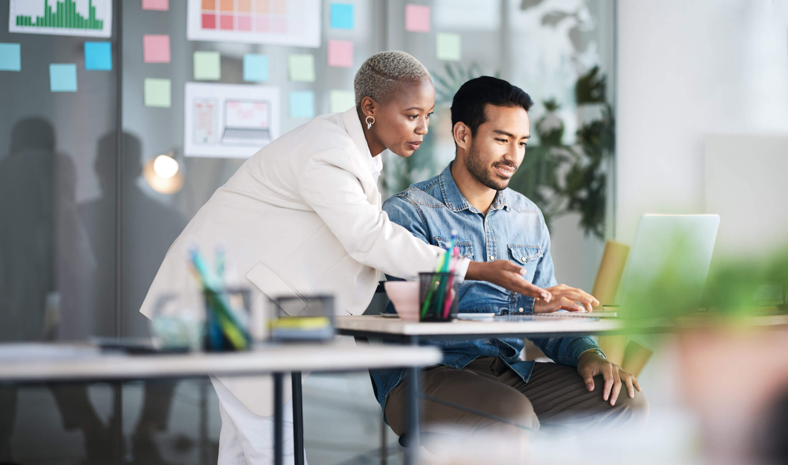 A woman in a white suit stands beside a man seated at a desk, pointing at his laptop screen. They appear to be discussing work in a modern office with colorful sticky notes on the glass wall behind them.