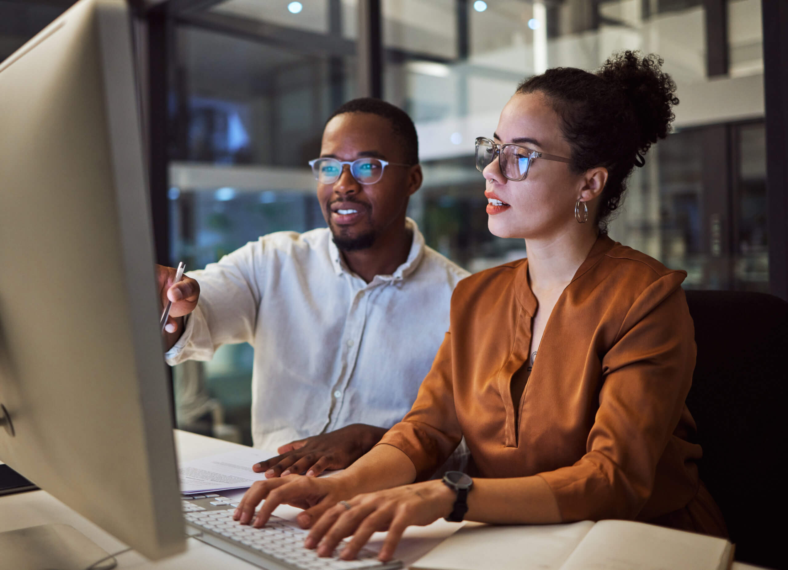 Two people wearing glasses sit at a desk, looking at a large computer monitor. One points at the screen while the other types, both appearing focused and engaged in a modern office setting.