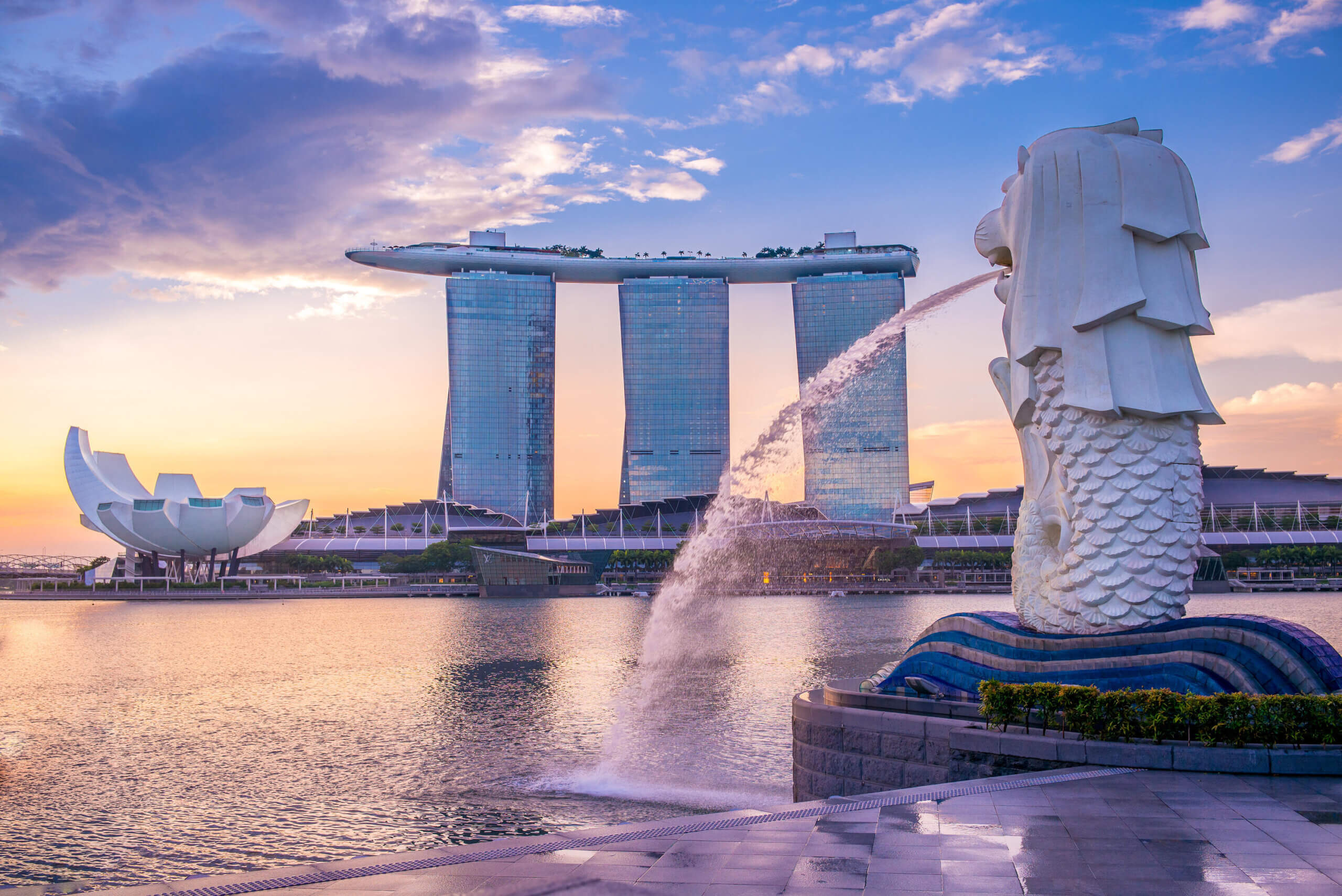 The Merlion statue spouts water into Marina Bay at sunset in Singapore, with the Marina Bay Sands hotel and the ArtScience Museum visible in the background under a partly cloudy sky.