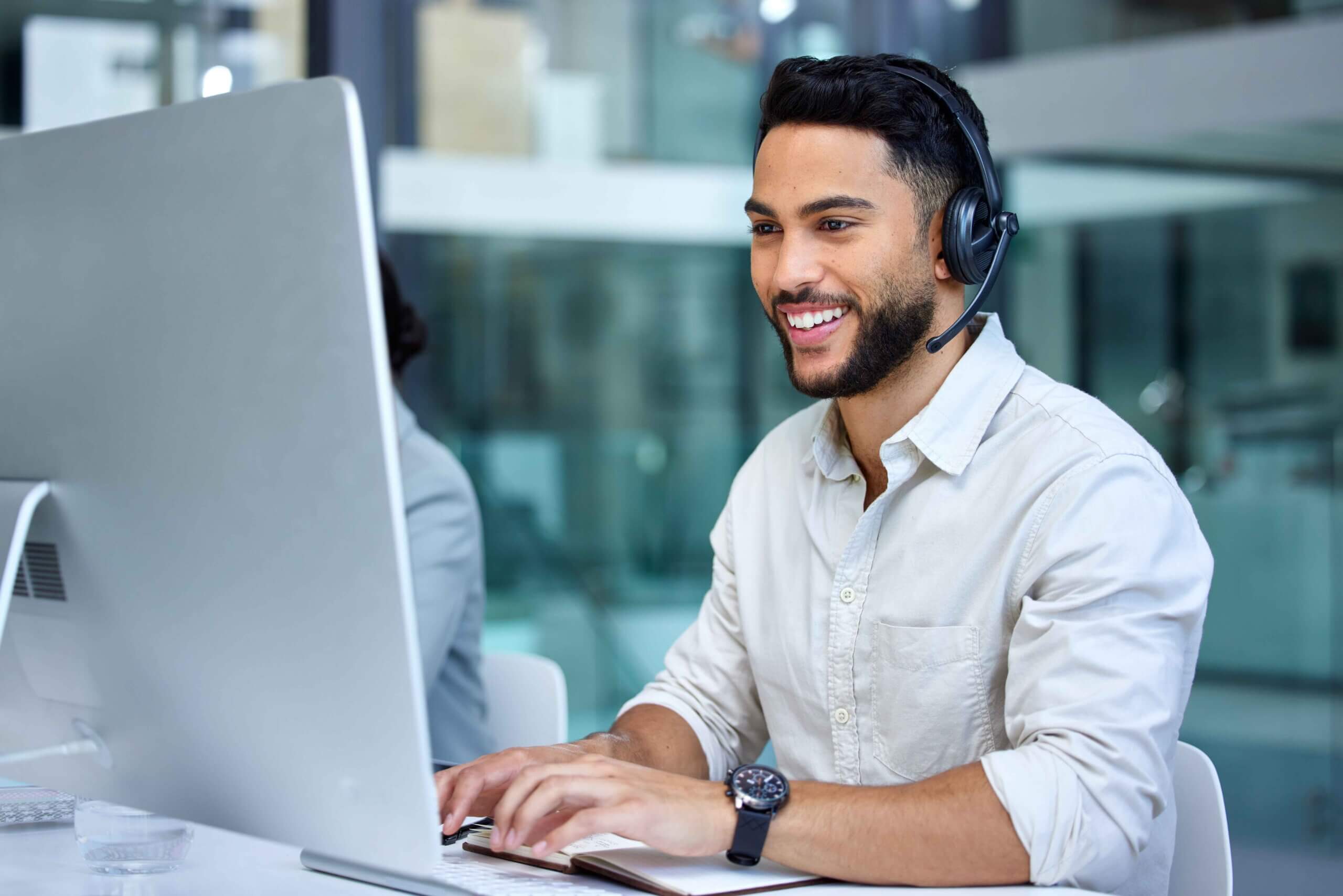 A man wearing a headset and a light-colored shirt sits at a desk, smiling while typing on a keyboard in front of a large computer monitor in a modern office setting.