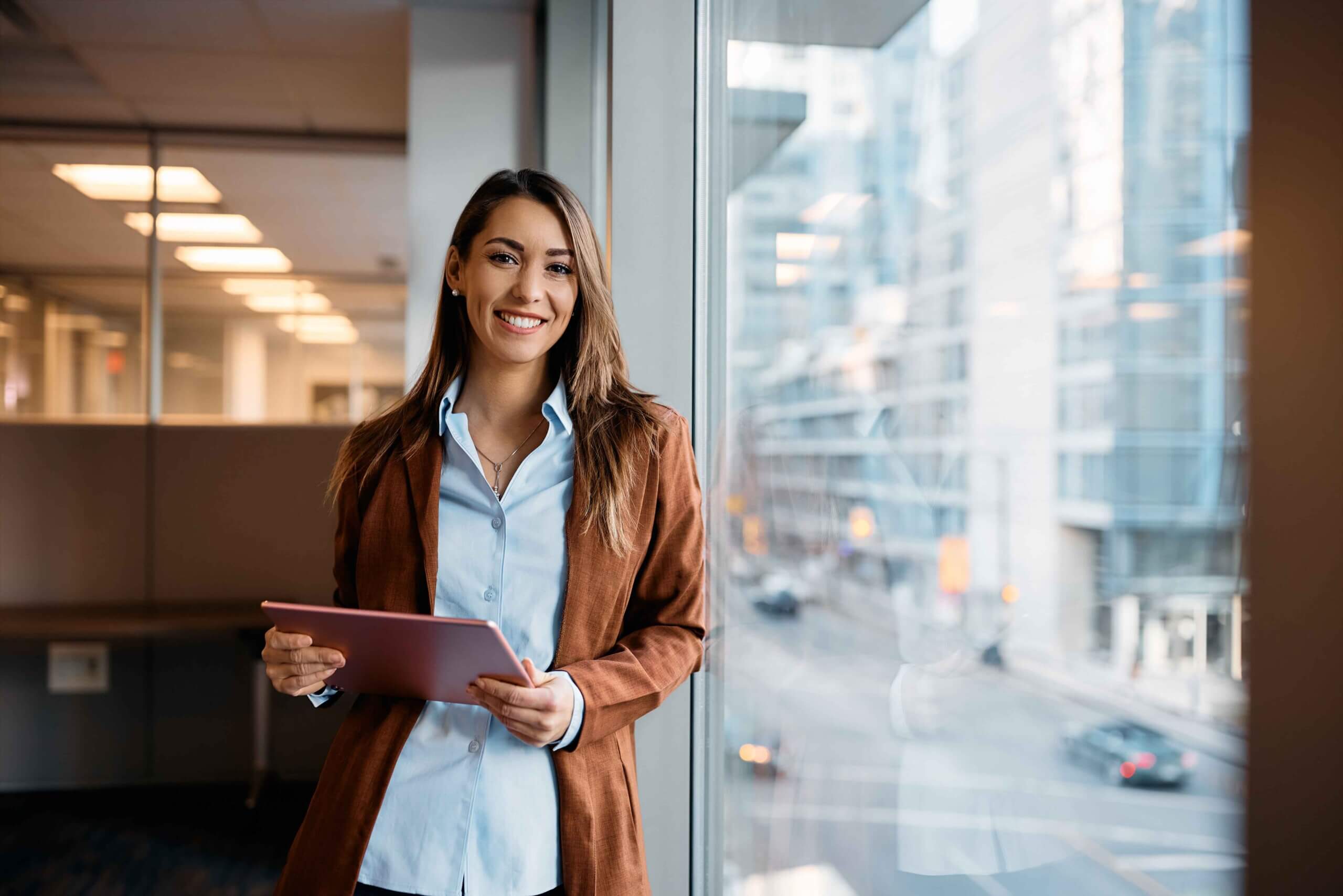 A woman in a brown blazer stands by a window in a modern office, smiling and holding a tablet. City buildings and a street with cars are visible outside through the glass.