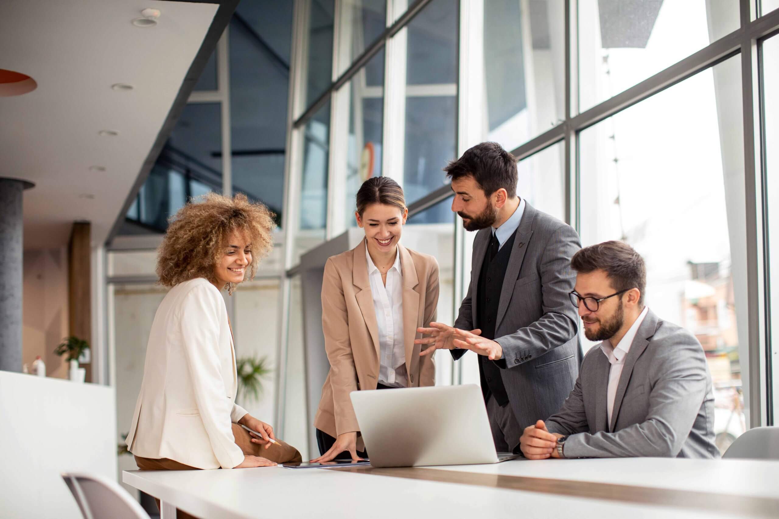 Four business professionals stand and sit around a table in a bright office, looking at a laptop and smiling as they discuss work together near large windows.