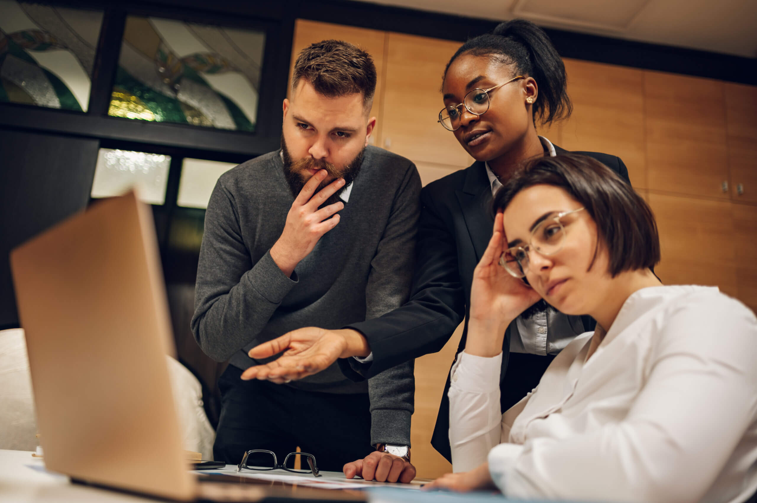 Three coworkers gathered around a desk, looking seriously at a laptop screen. One gestures while the other two appear concerned and thoughtful, suggesting a tense or challenging work situation.