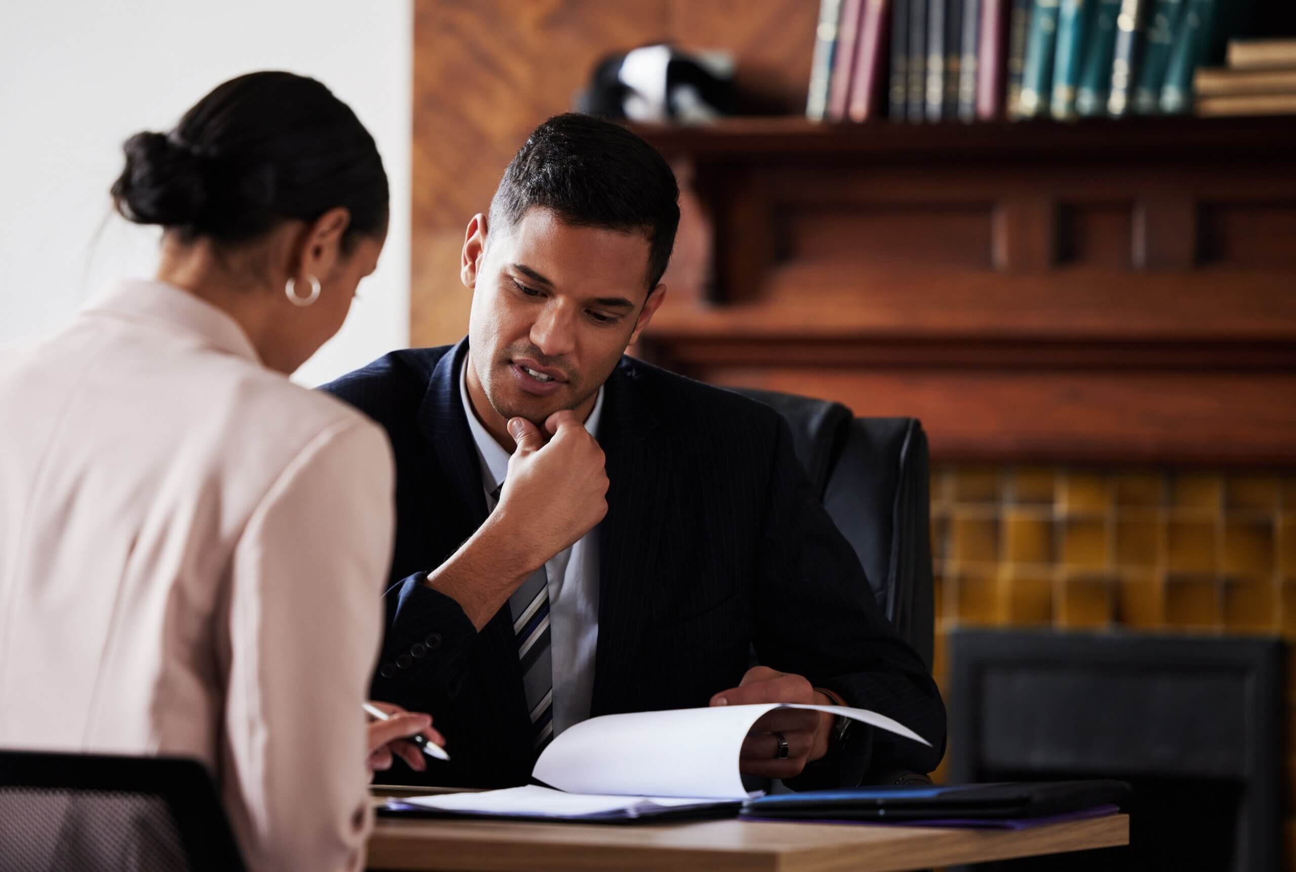 Two people in business attire sit at a desk in an office, discussing documents. One person is holding papers while the other listens attentively. Books and wooden shelves are in the background.