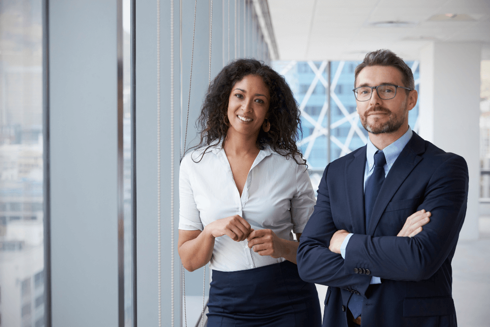 Two business professionals, a woman and a man, stand in a modern office by large windows. The woman is smiling with her hands clasped, and the man is wearing glasses and has his arms crossed, both dressed in business attire.