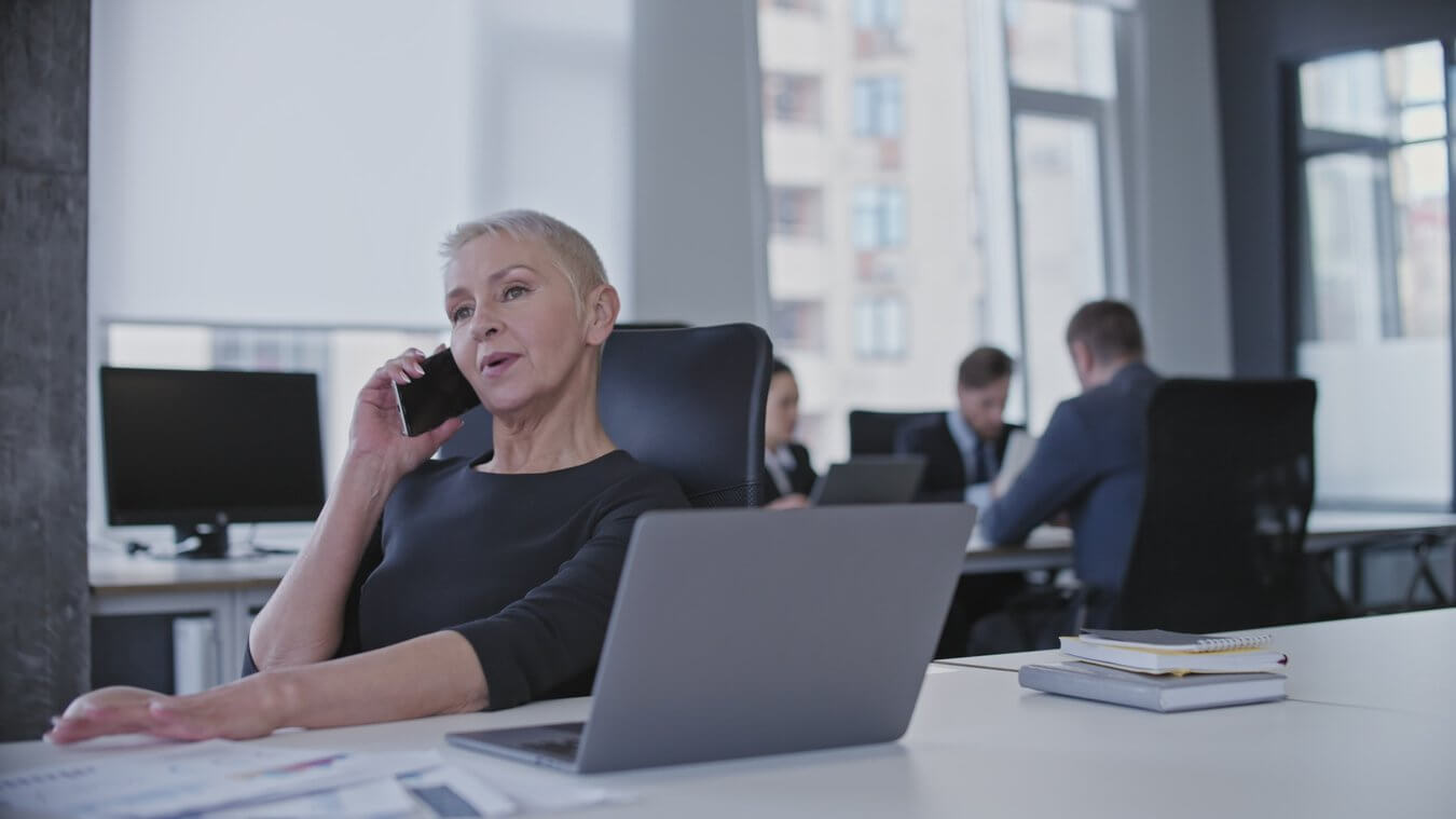 A woman with short gray hair sits at a desk in an office, talking on a smartphone. A laptop and some notebooks are on her desk. In the background, four people are having a meeting at another table.